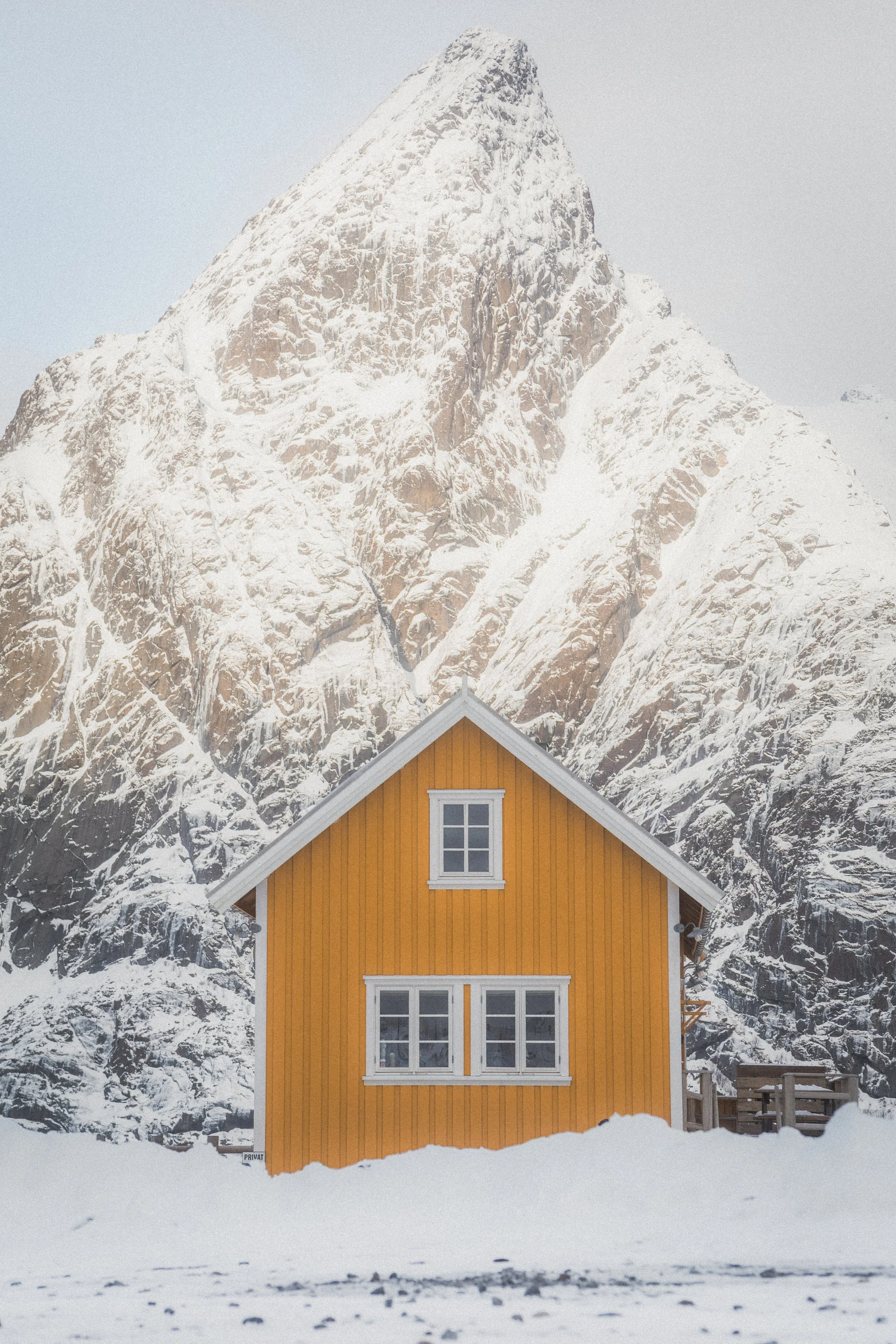 A yellow wooden house with white window frames in a snowy landscape, with a large, snow-covered mountain in the background.