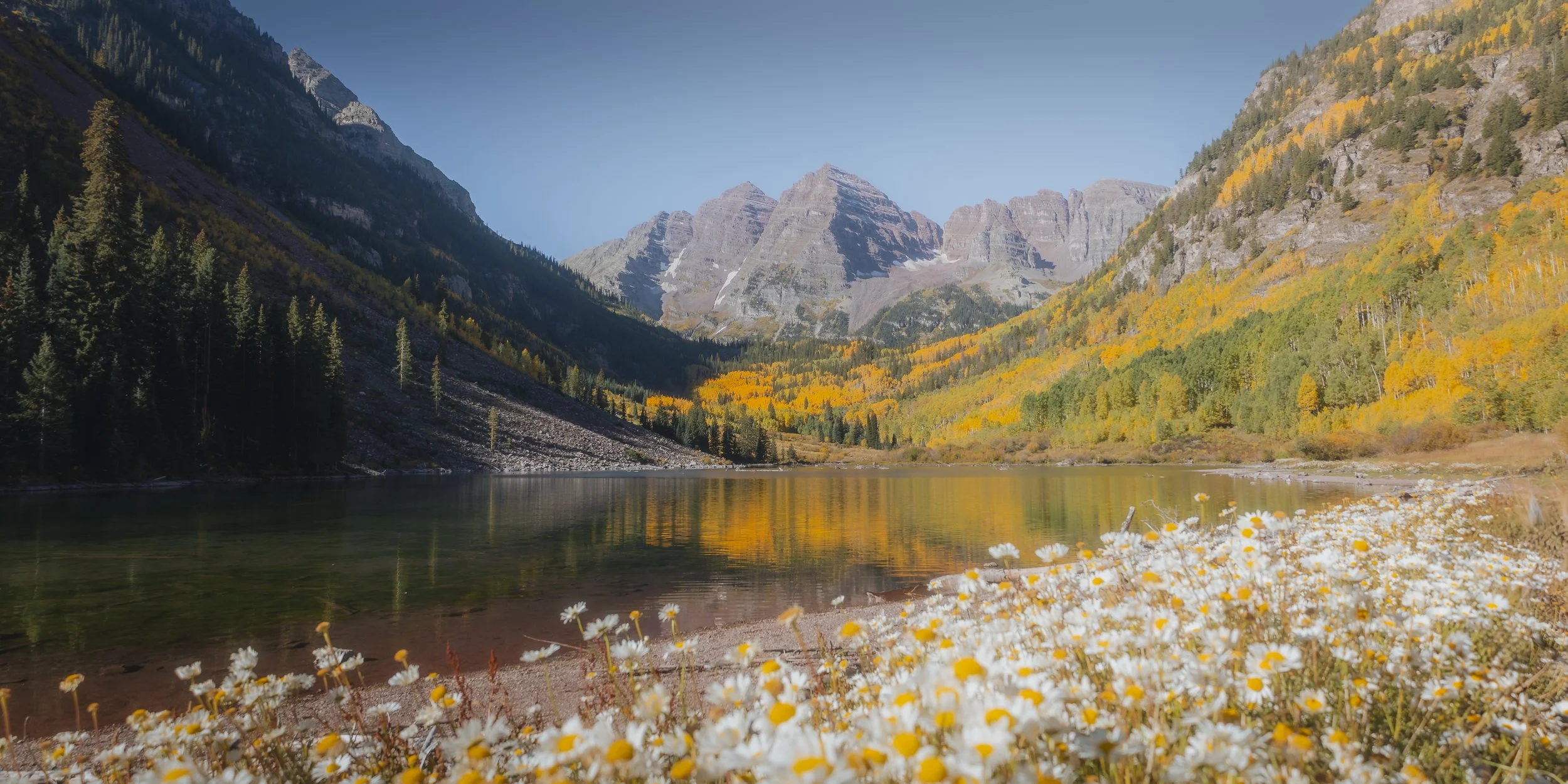 A picturesque mountain landscape with a lake in the foreground, surrounded by trees with autumn foliage and snow-capped peaks in the distance.