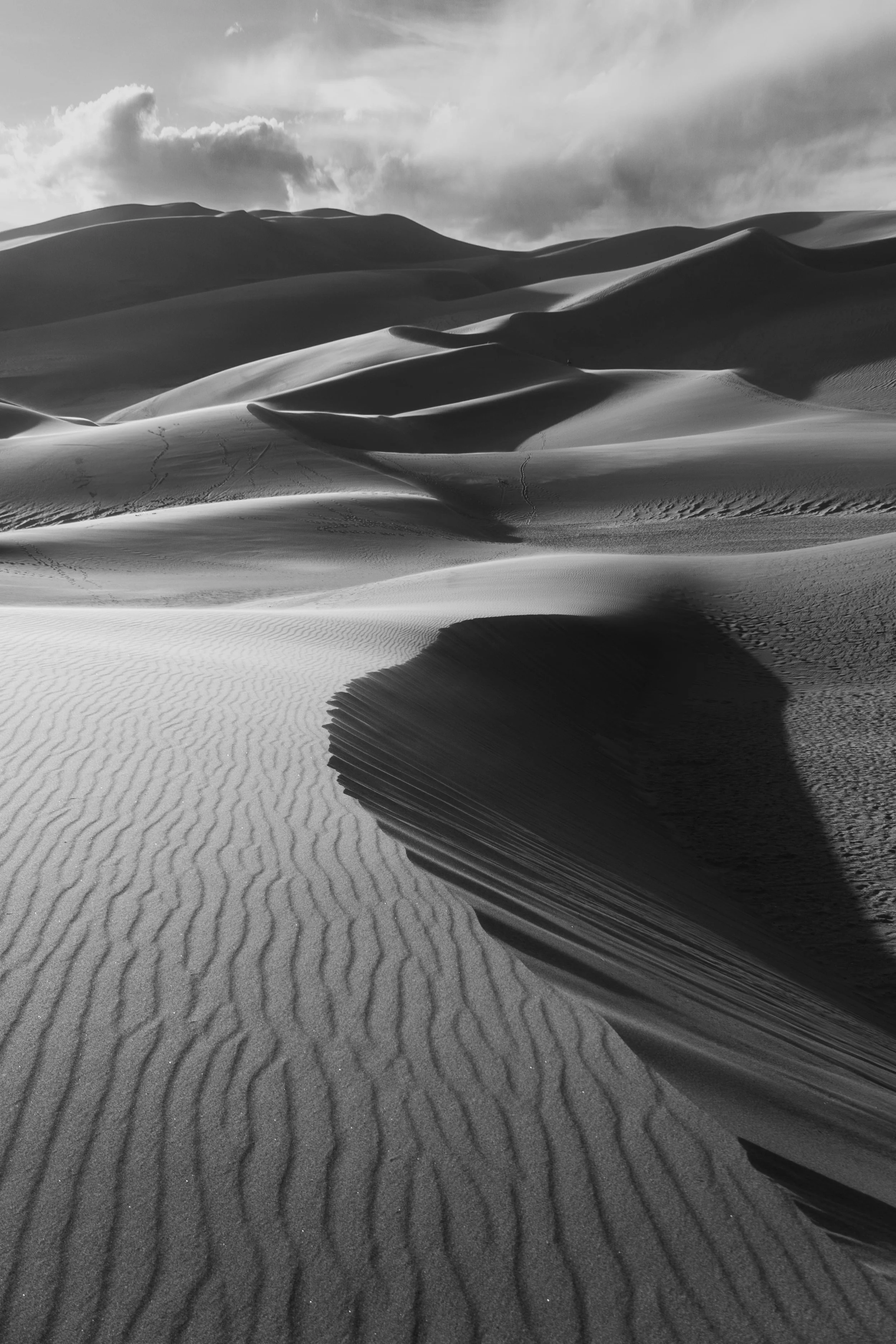 Black and white photo of desert sand dunes with wind-shaped ridges and a cloudy sky.