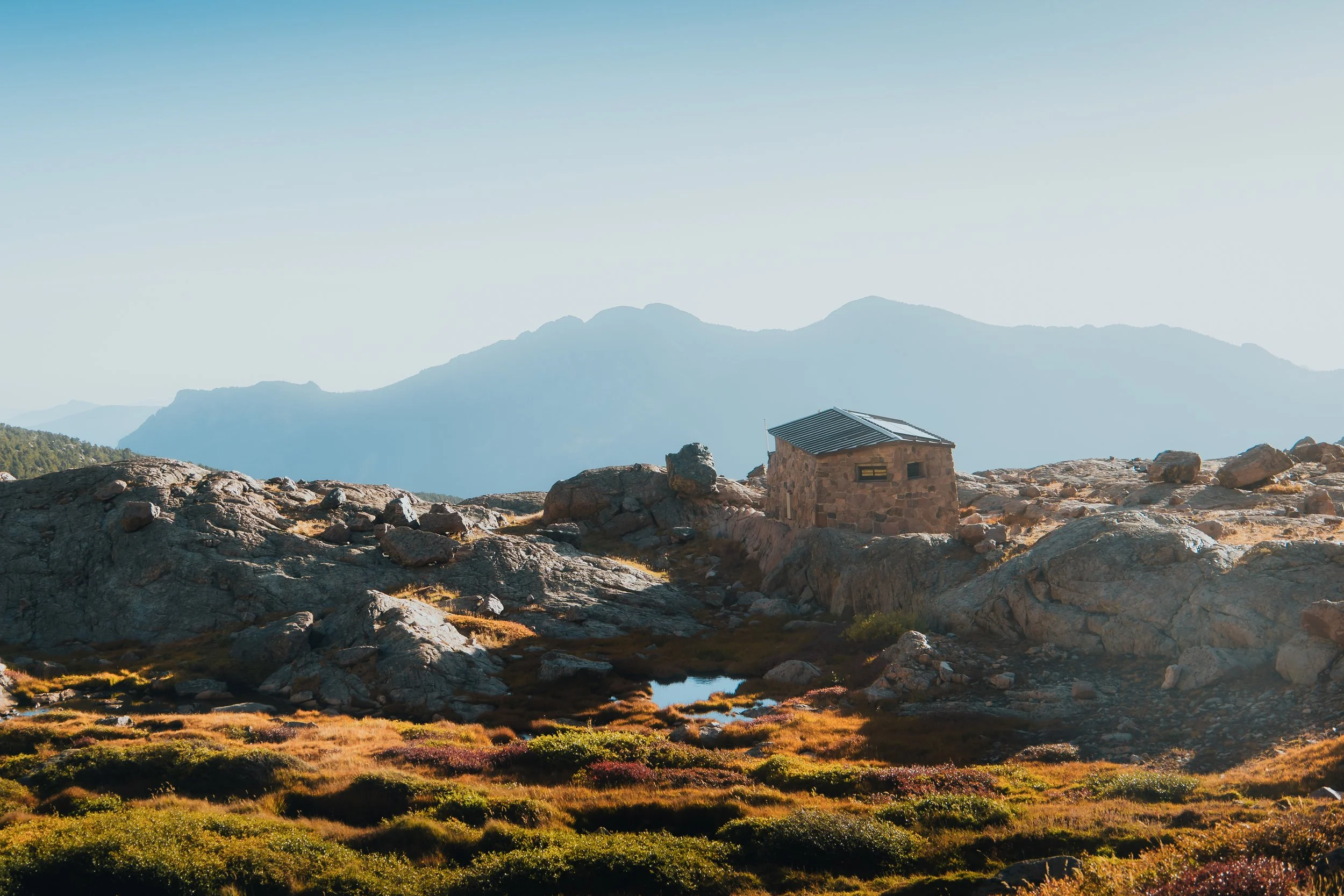 A small stone cabin with a sloped roof in a rocky mountain landscape with colorful ground vegetation, distant mountains, and a clear blue sky.