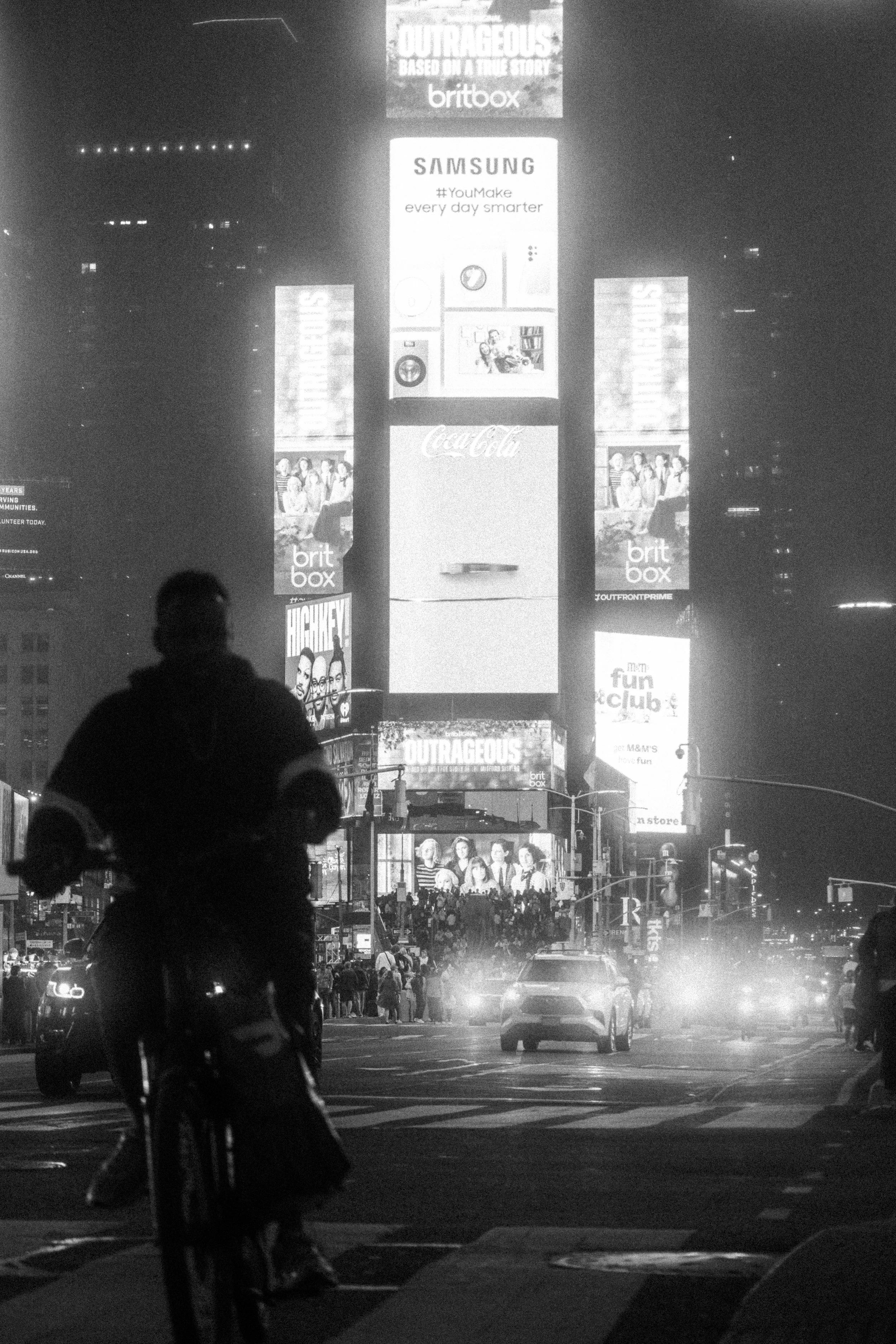 A black and white photo of Times Square at night, featuring illuminated billboards, a cyclist in the foreground, and a crowd of people crossing the street.
