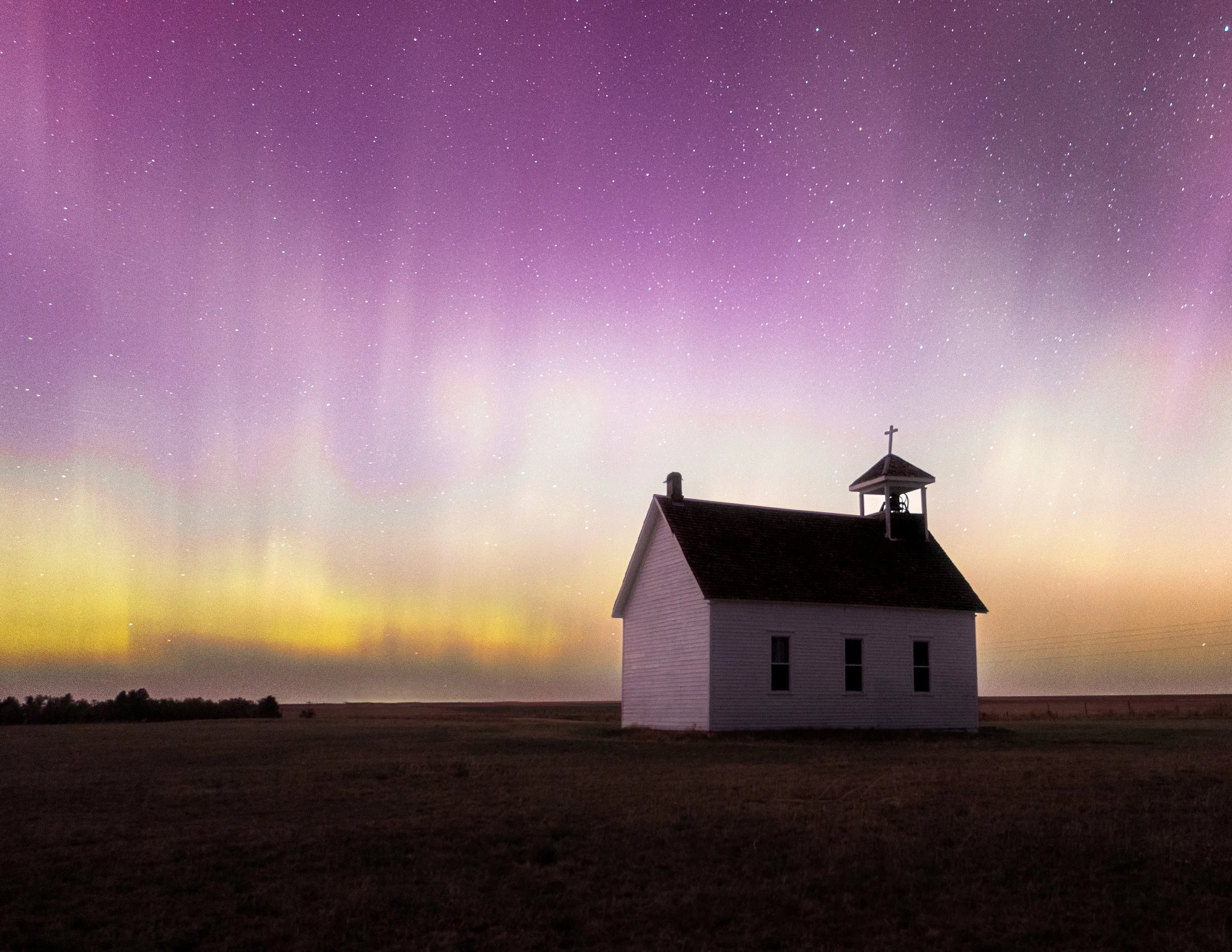 A small white church with a steeple and cross in a vast open field under a colorful starry night sky with visible aurora borealis.