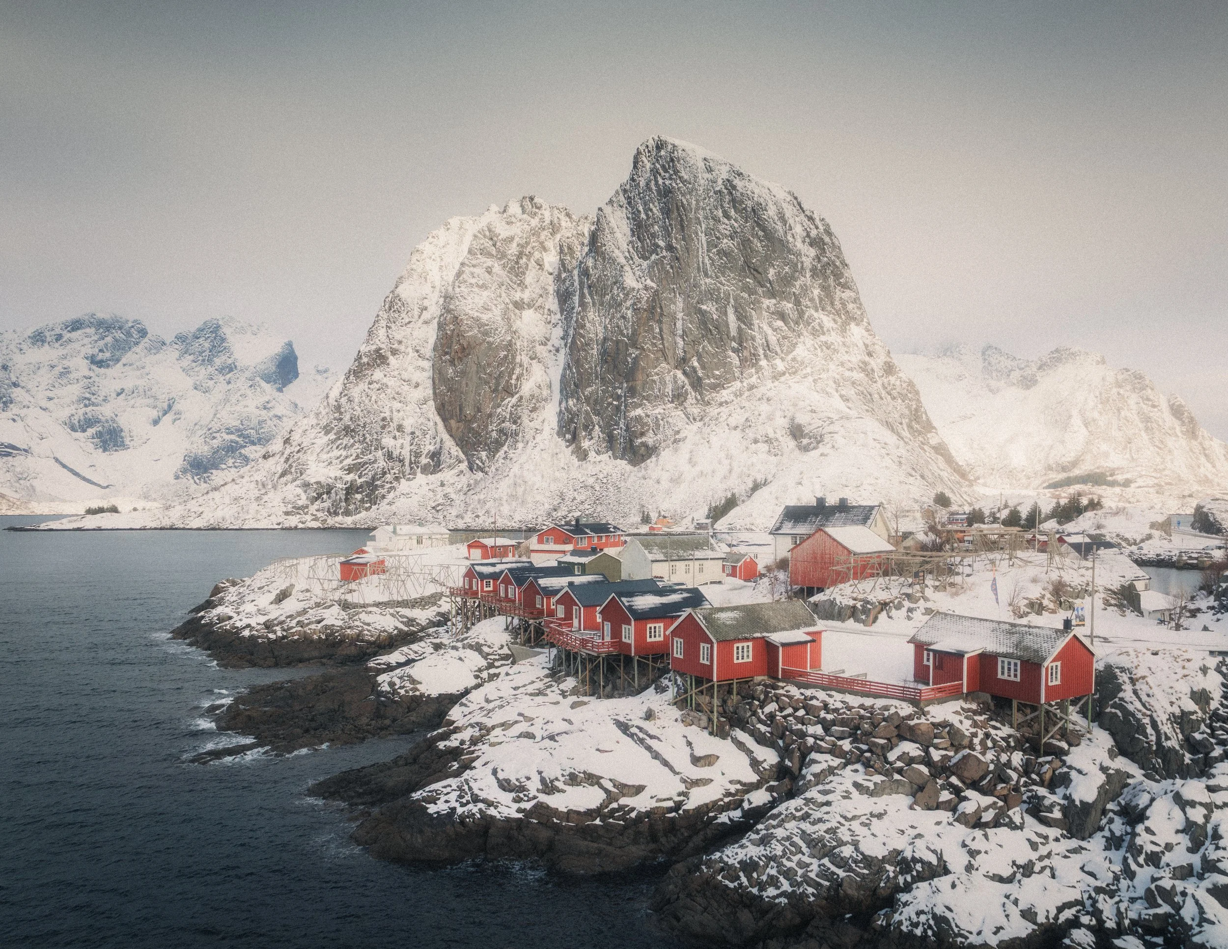 Snow-covered village with red houses on rocky shoreline, mountainous snow-covered landscape in the background, over water.