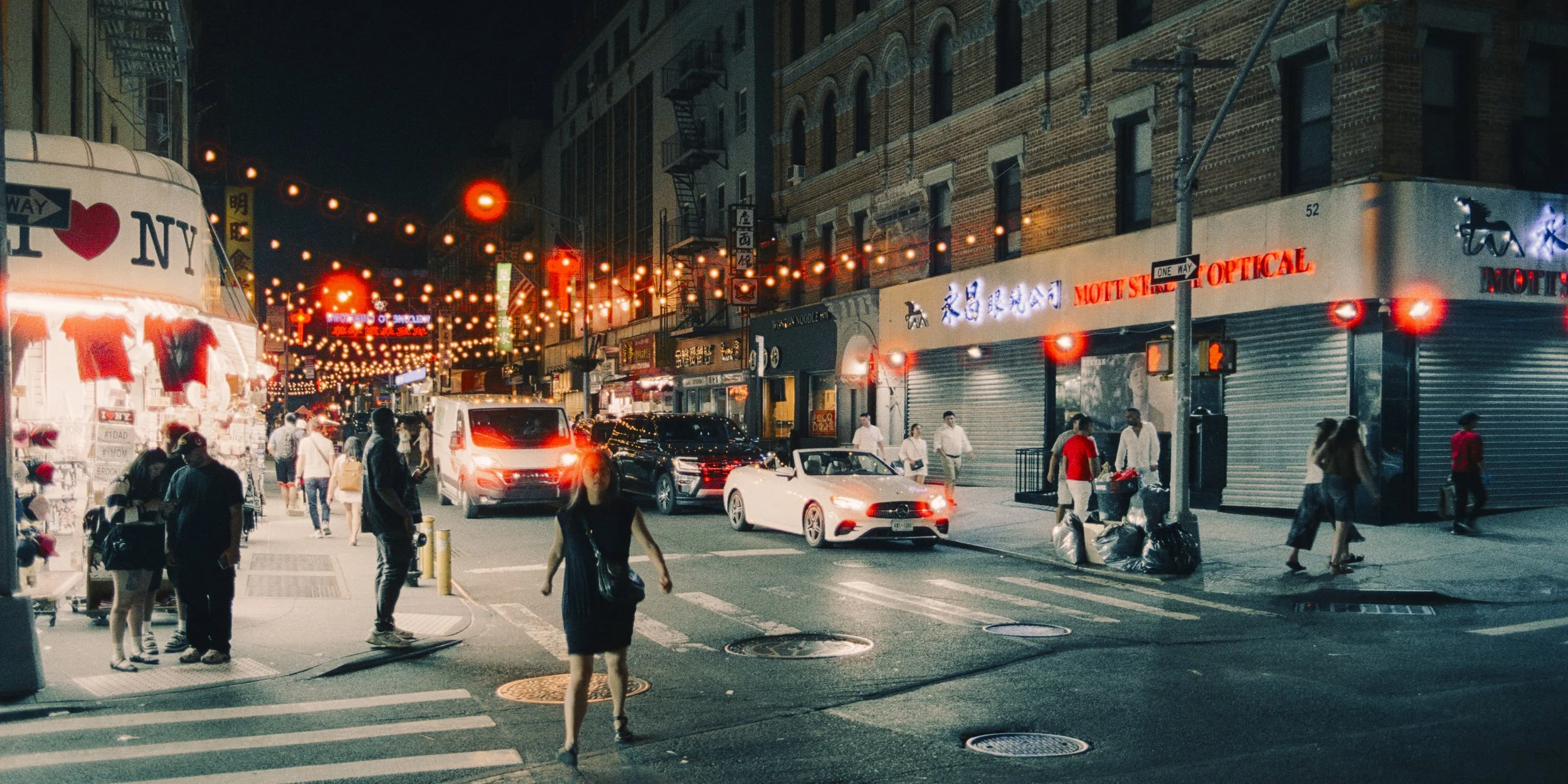Night view of a busy city street with pedestrians crossing and cars driving, illuminated by string lights and storefront signs.
