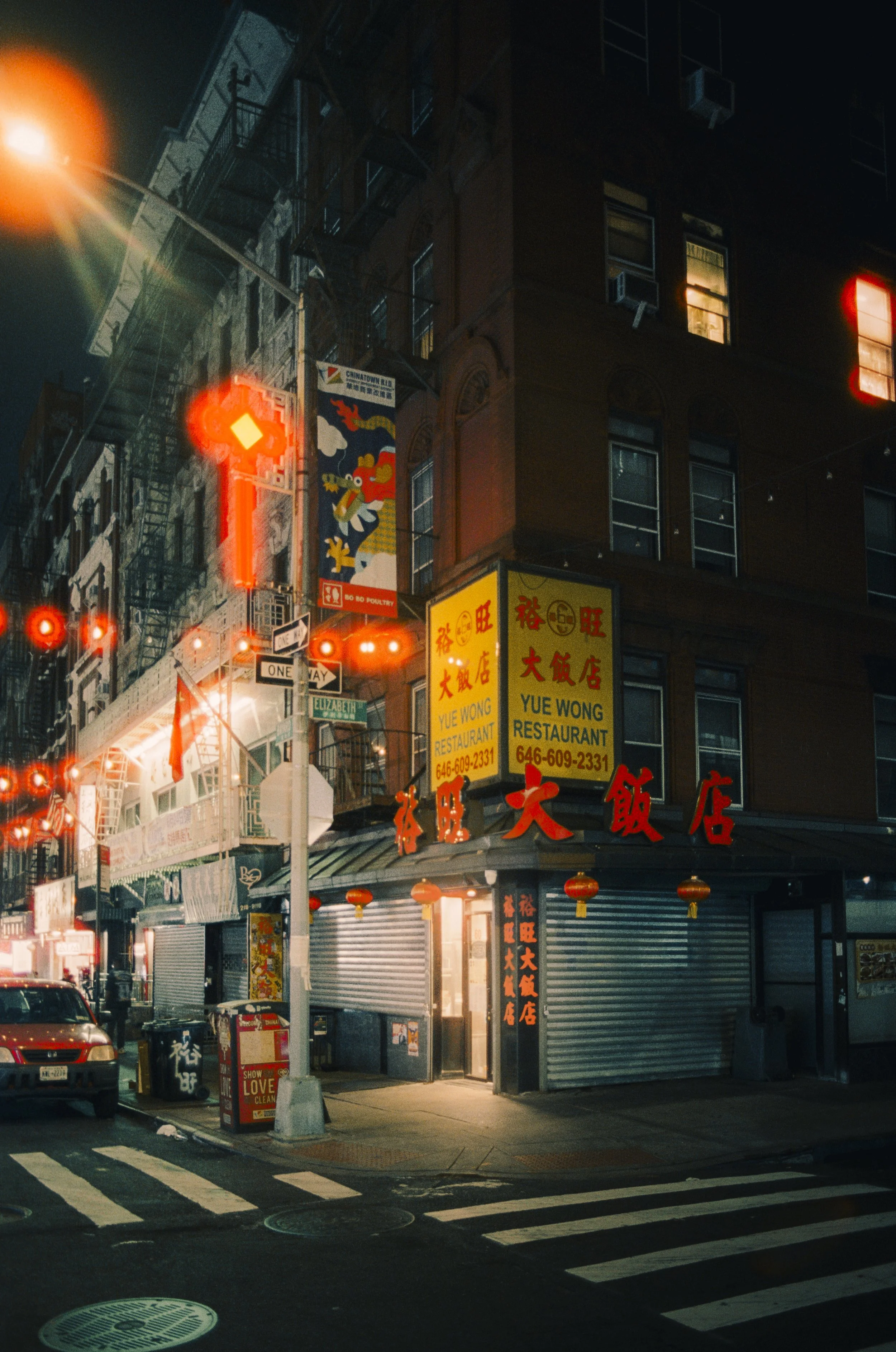 Nighttime street scene with illuminated signs of a Chinese restaurant, Yue Wong Restaurant, and decorative red lanterns, with a crosswalk and cars in the foreground.