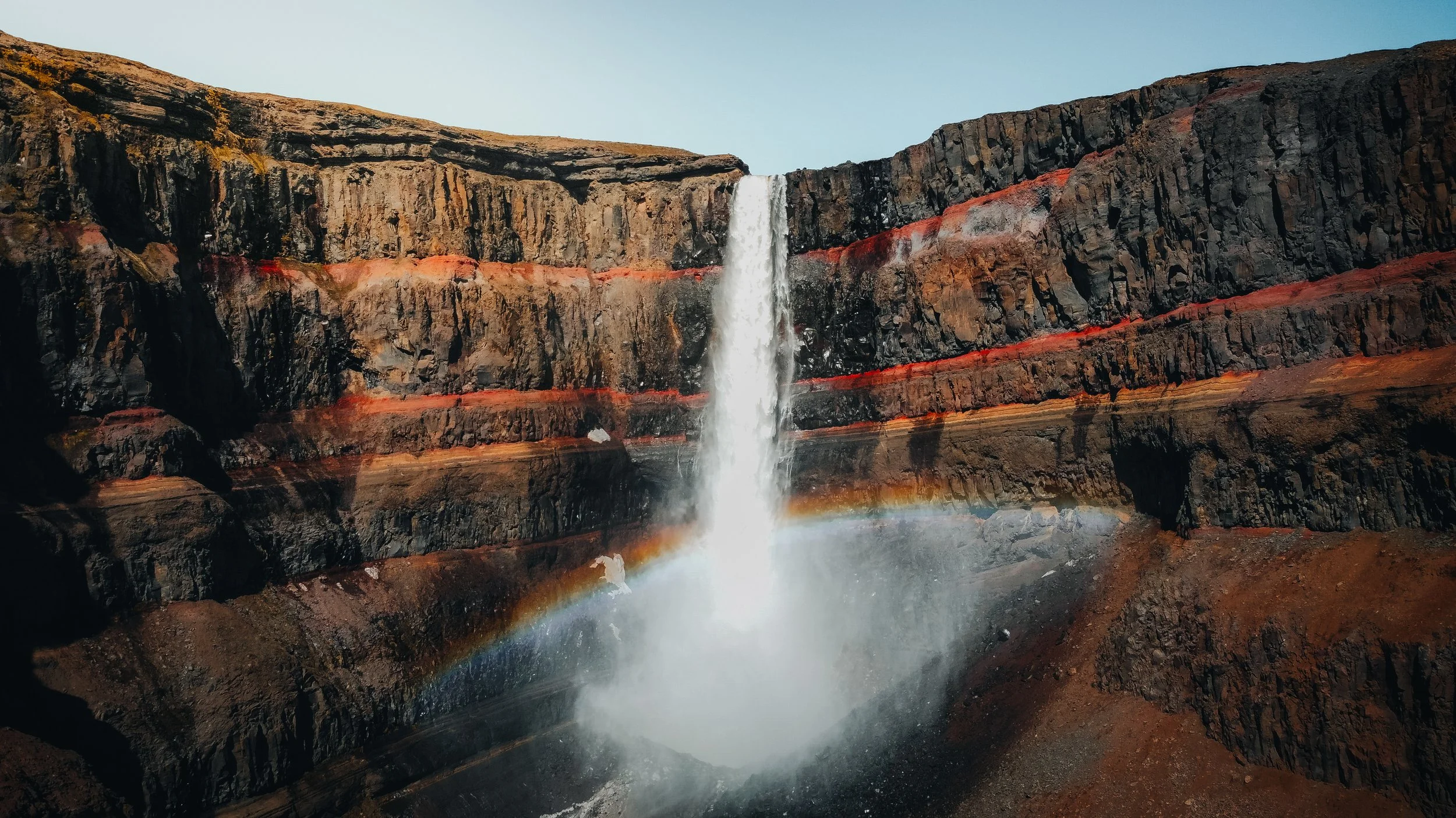 Waterfall cascading down a tall, rugged cliff with stratified rock layers, some red and orange, and a rainbow forming at the base
