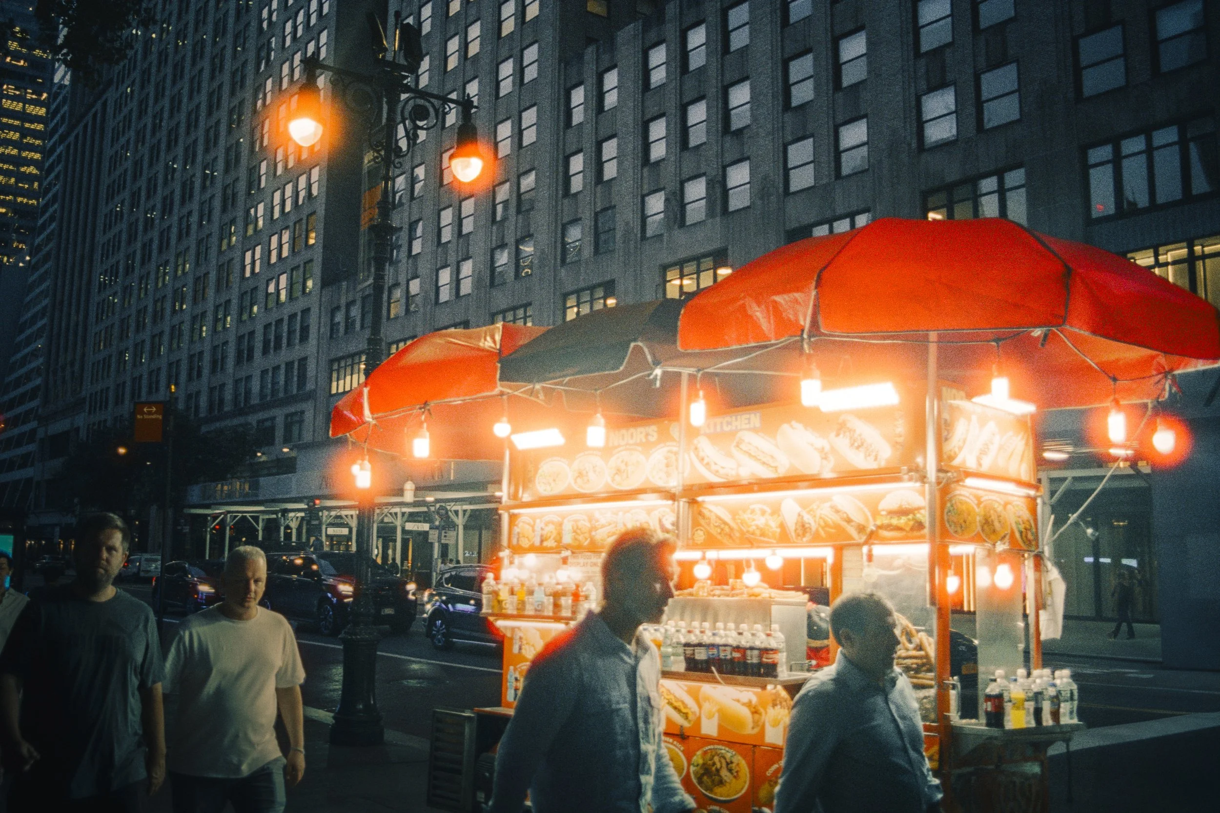 A city street scene at dusk with a bright orange hot dog stand illuminated by red and warm lights. Pedestrians are walking by, and tall buildings with many windows are in the background. The hot dog stand has images of hot dogs and food items on its 