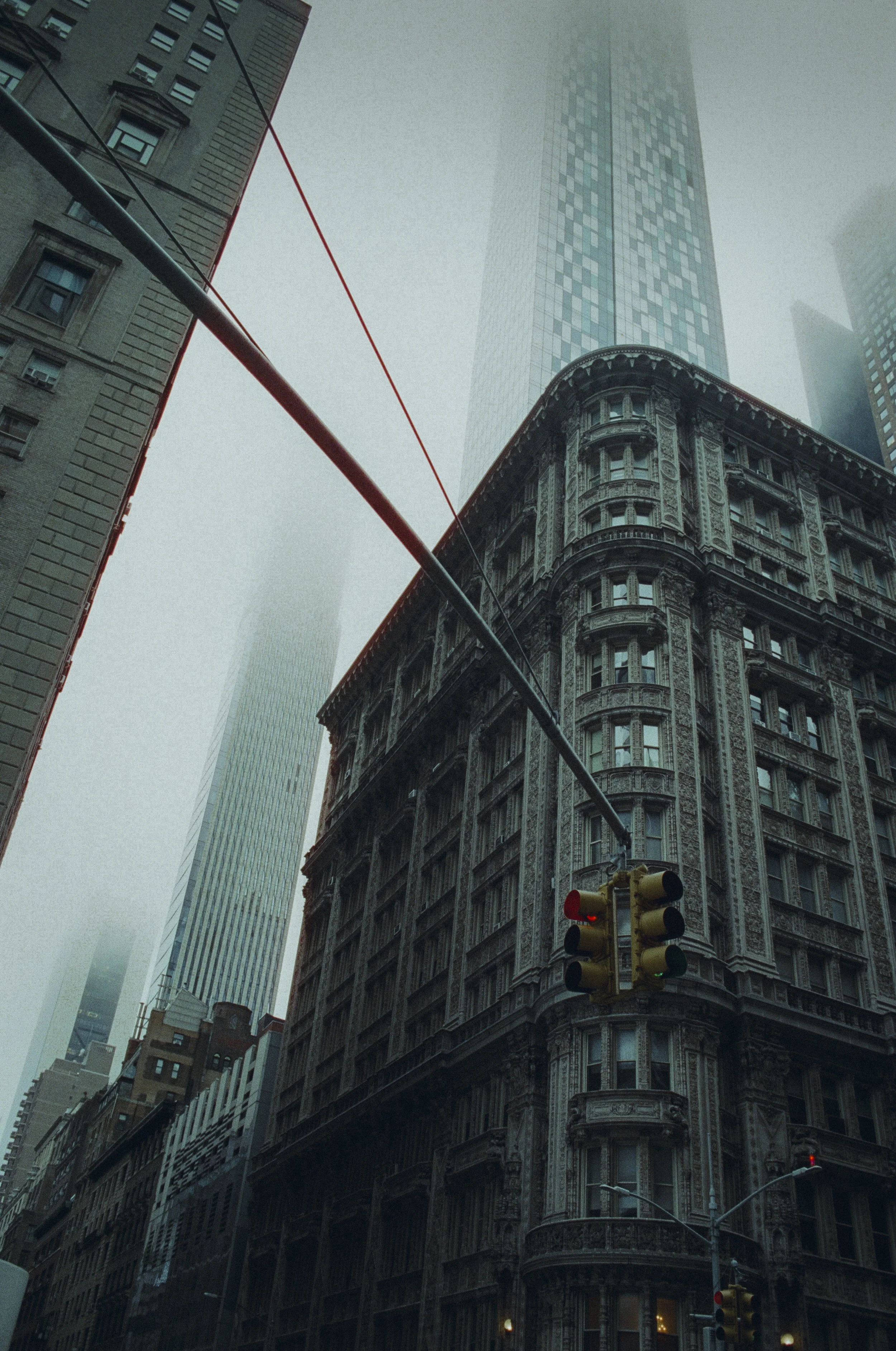 A city street scene with tall buildings, including a historic ornate building in the foreground, high-rise skyscrapers in the background, and traffic lights hanging at the intersection on a foggy or misty day.