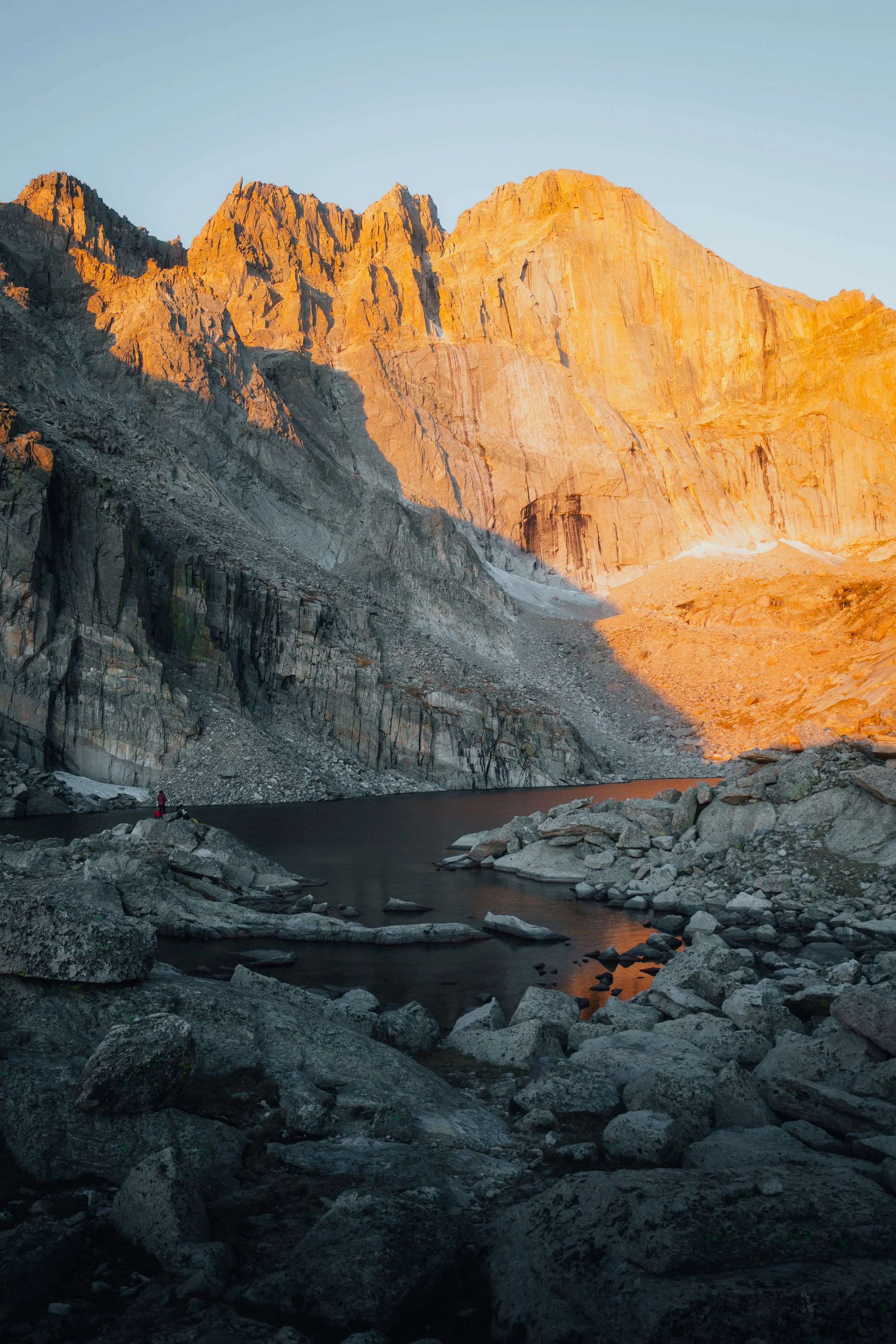 A mountain landscape at sunset with rocky peaks illuminated in orange light, a small pool of water in the foreground, and a person standing on rocks near the water.