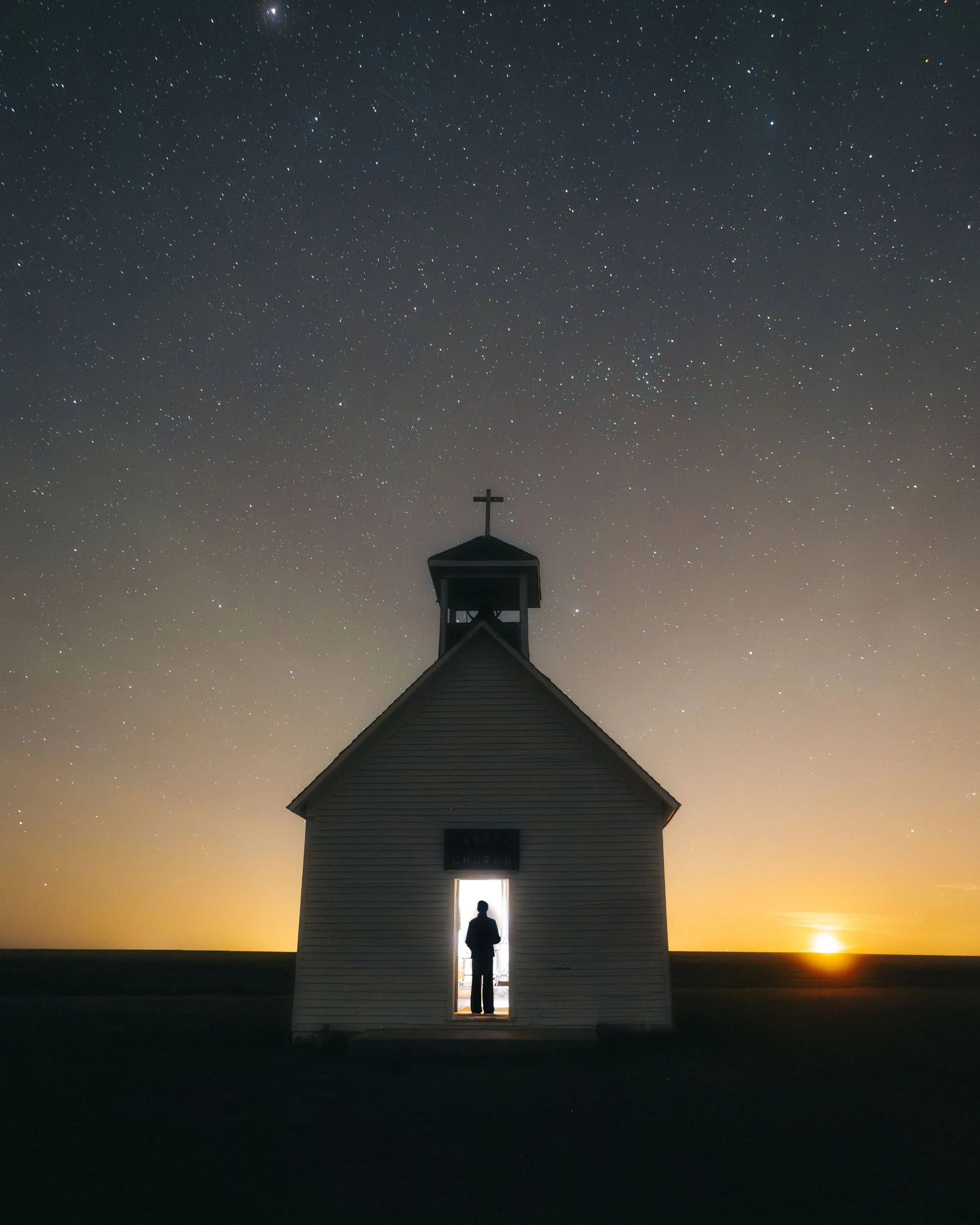 A small church silhouette at sunset or dusk with a person standing in the doorway, under a starry night sky.