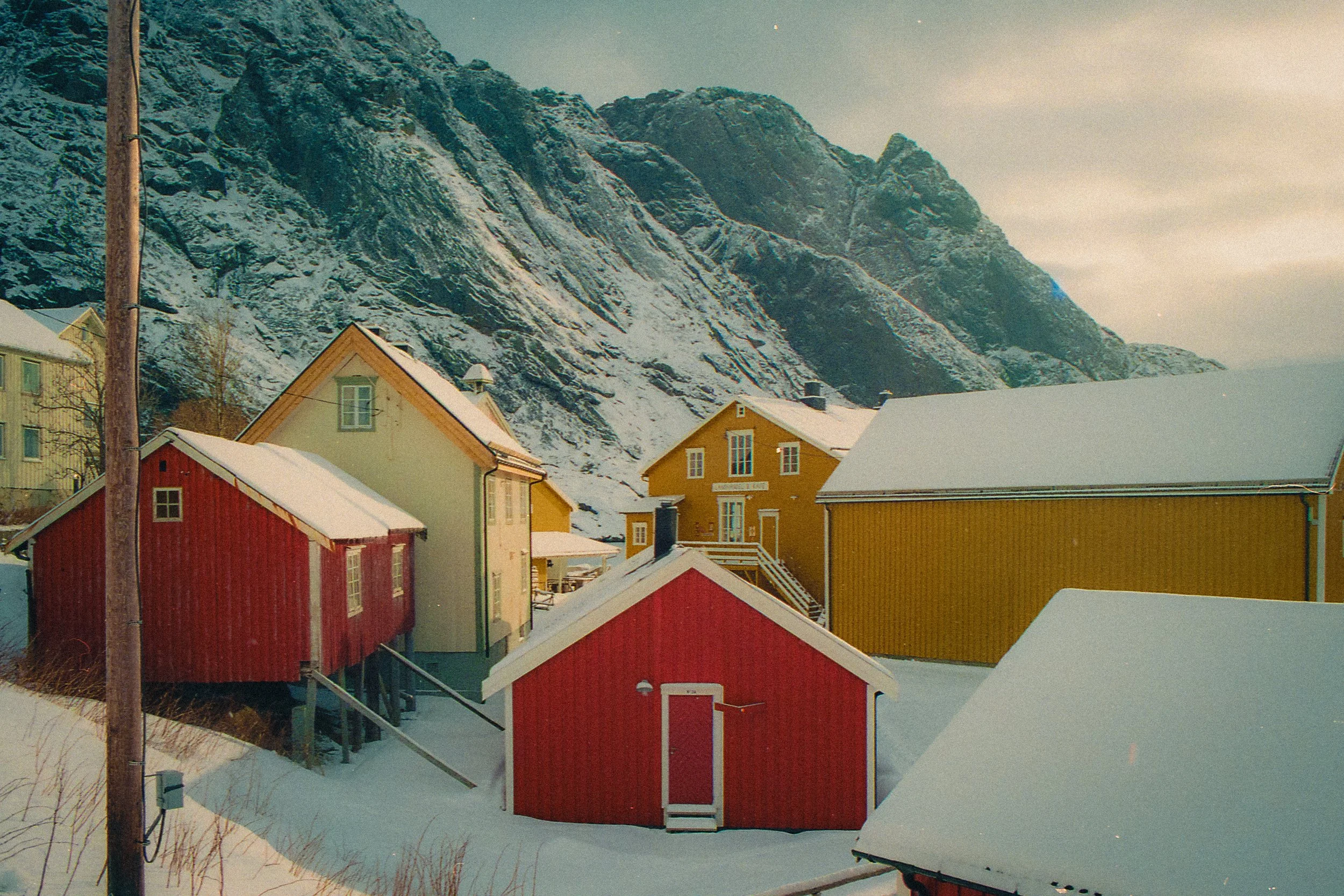 Colorful houses with snow-covered roofs in front of a mountain range with snow, under a cloudy sky.