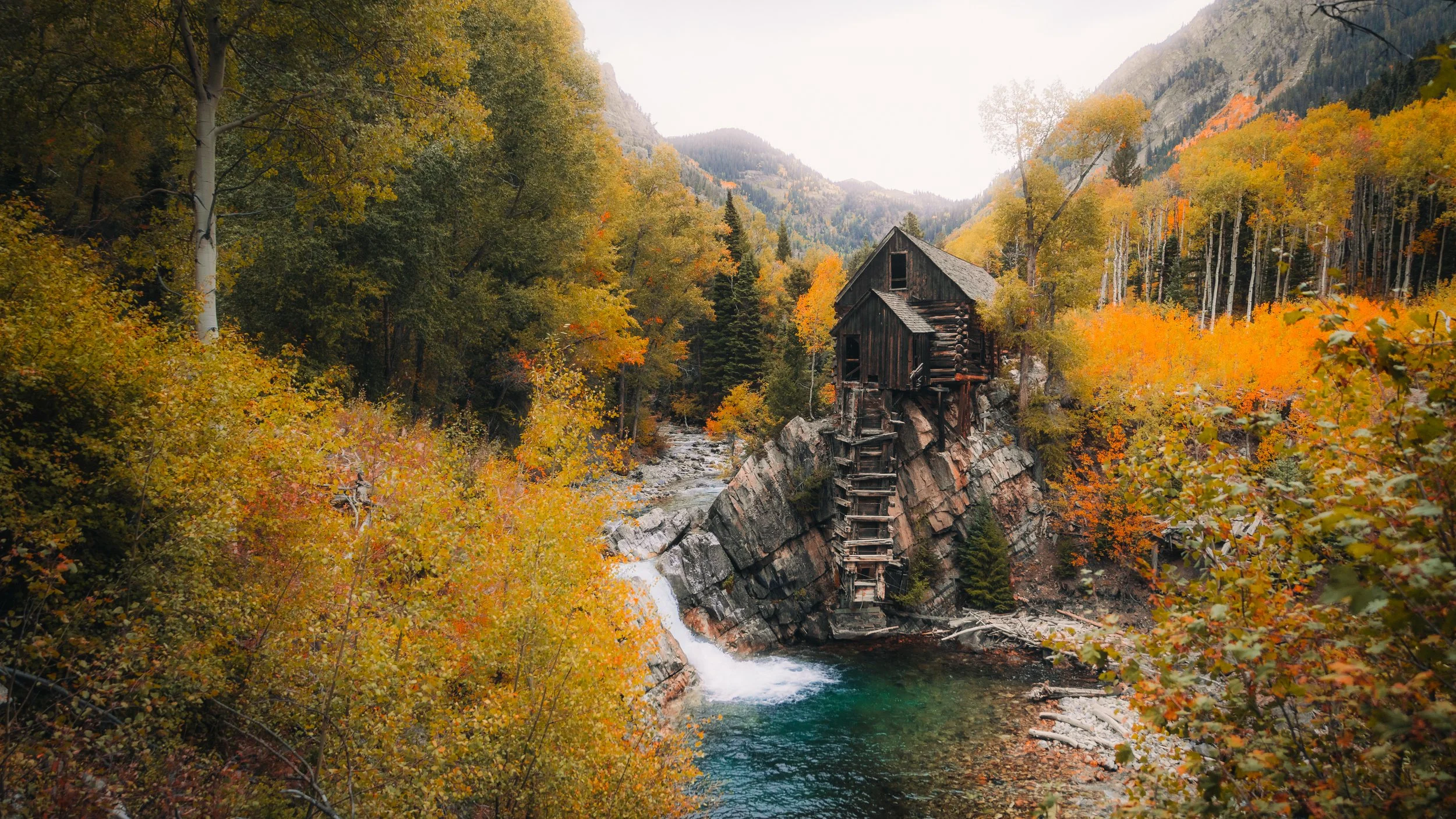An abandoned wooden mill on a rocky outcrop over a river, surrounded by autumn trees in a mountainous landscape.