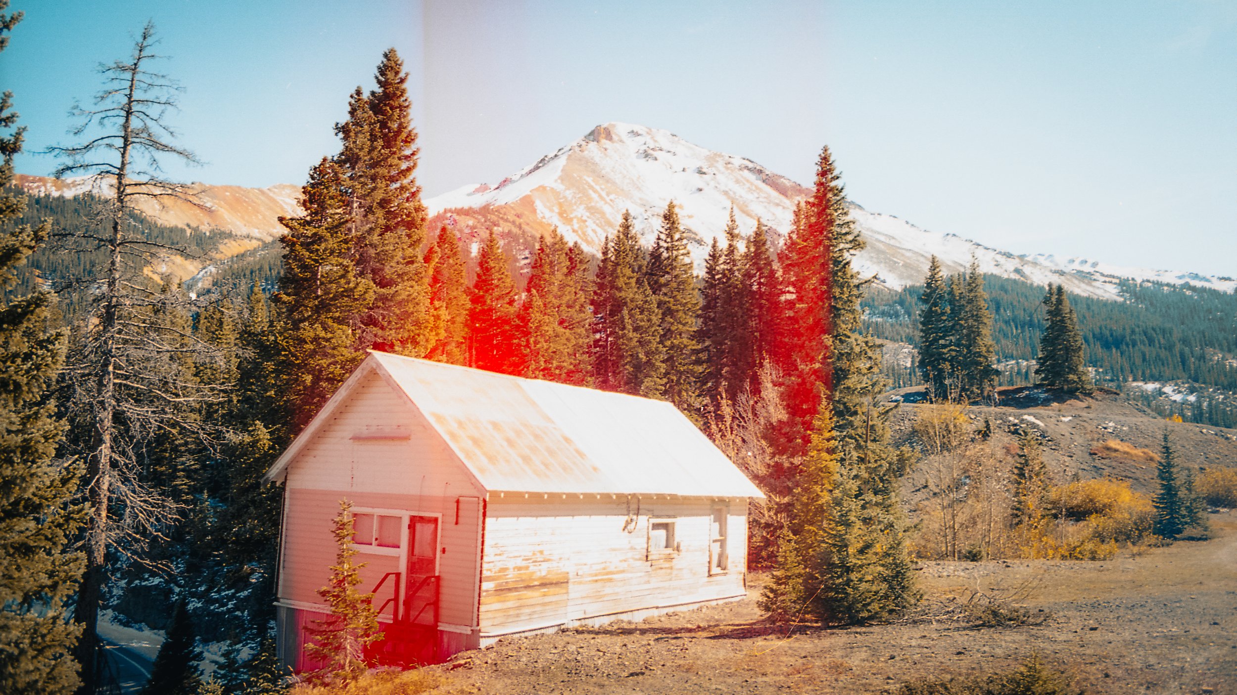 A small white wooden house with a rusted roof is situated among tall green pine trees in a mountainous landscape with snow-capped peaks in the background.