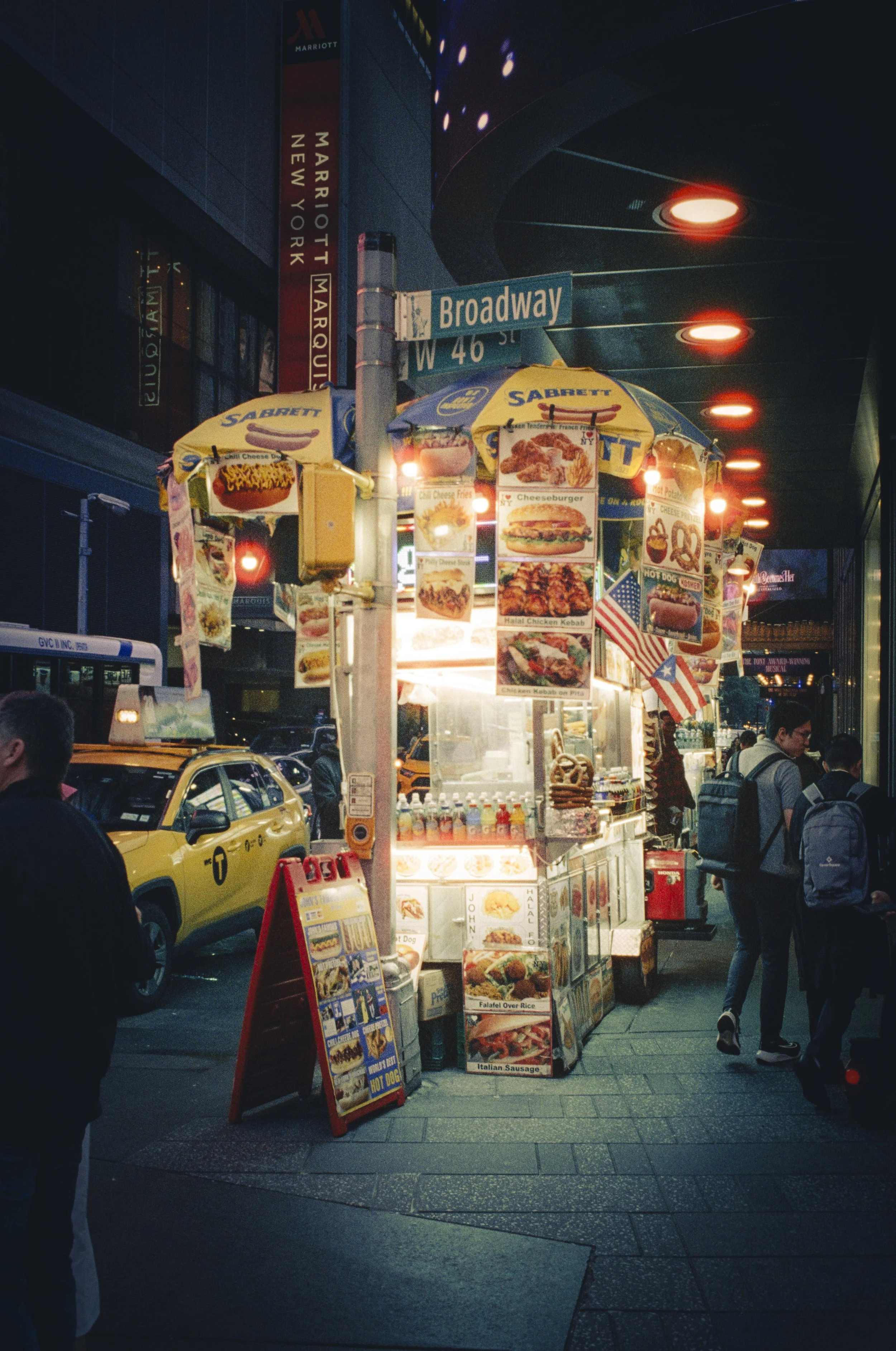 A street food stand with illuminated signs offering burgers, hot dogs, chicken, and fries in New York City at night, with pedestrians and yellow taxis nearby.