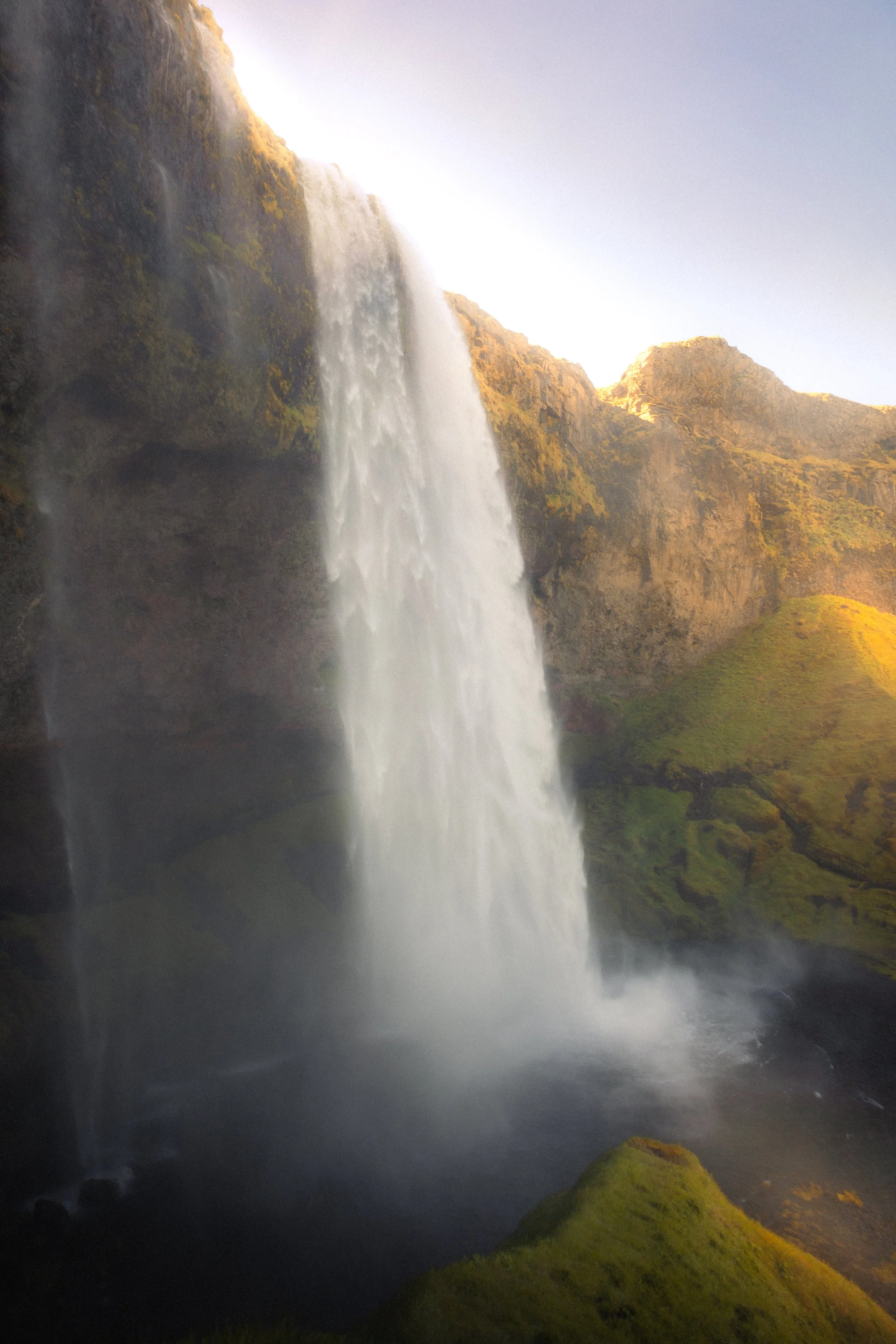 A tall waterfall cascading down a cliff with moss-covered rocks and lush green hills in the background.