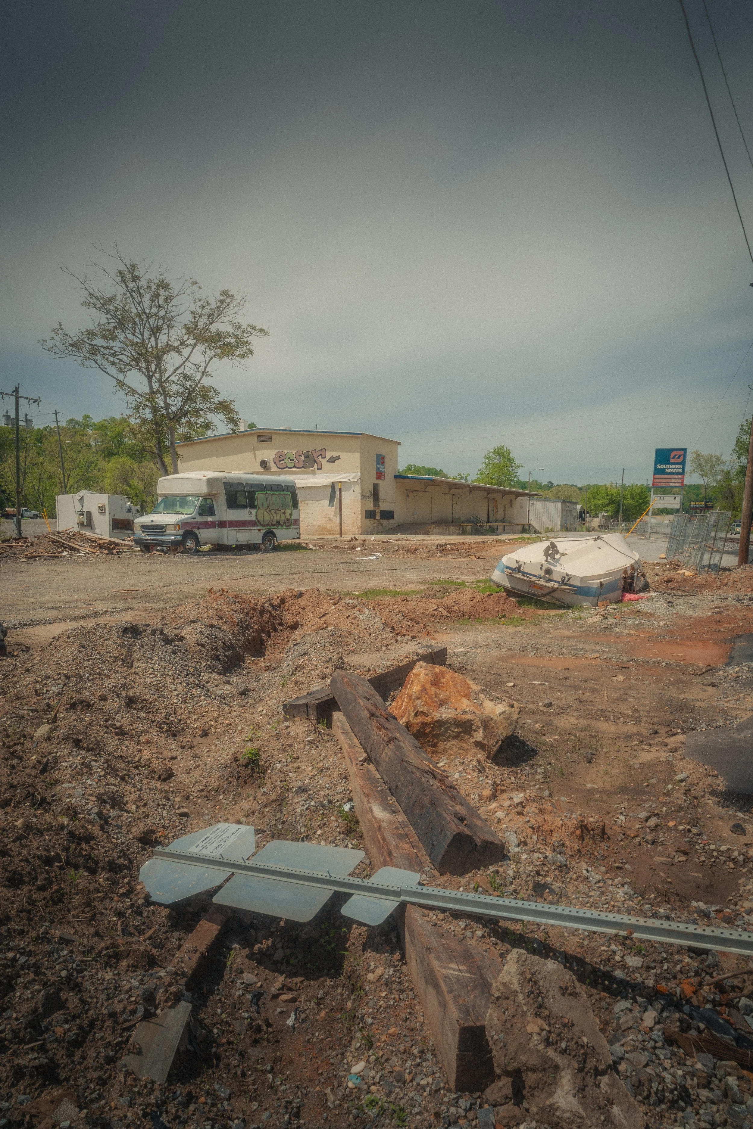A disturbed dirt lot with scattered debris, including a fallen wooden beam, a metal vent pipe, and rocks. In the background, there is an abandoned building with graffiti, a boat on the ground, and a white van parked nearby. The sky is overcast.