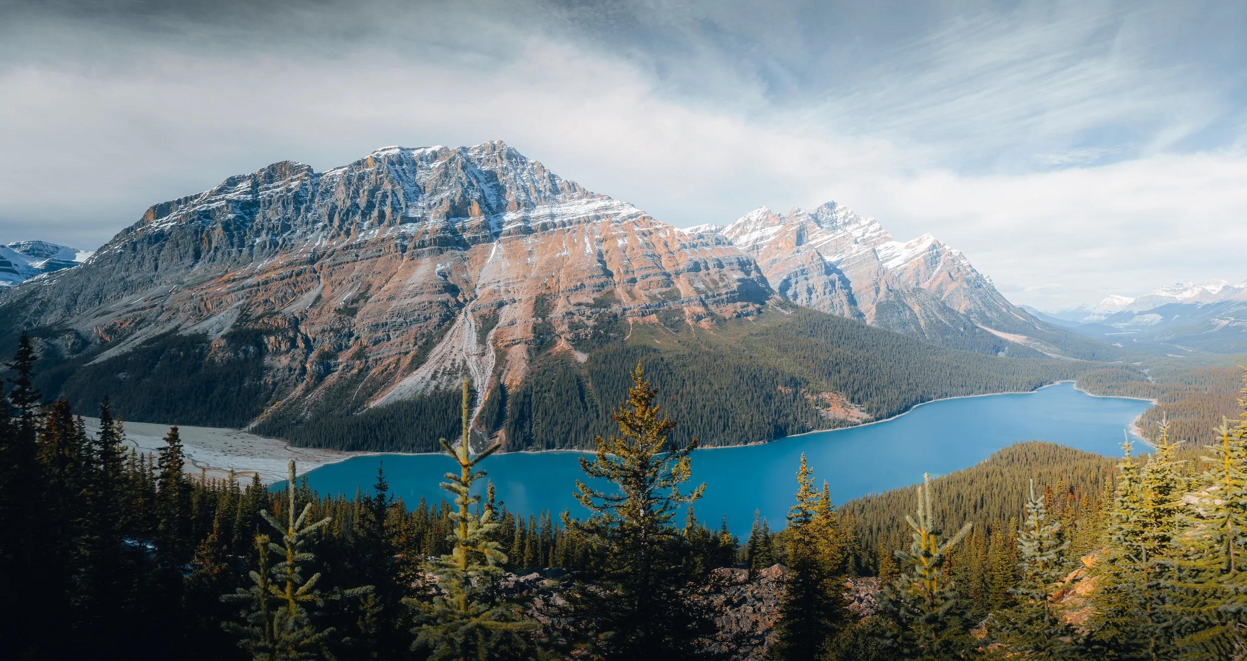 A scenic mountain landscape with a large lake in the foreground, surrounded by dense evergreen trees and snow-capped peaks under a partly cloudy sky.