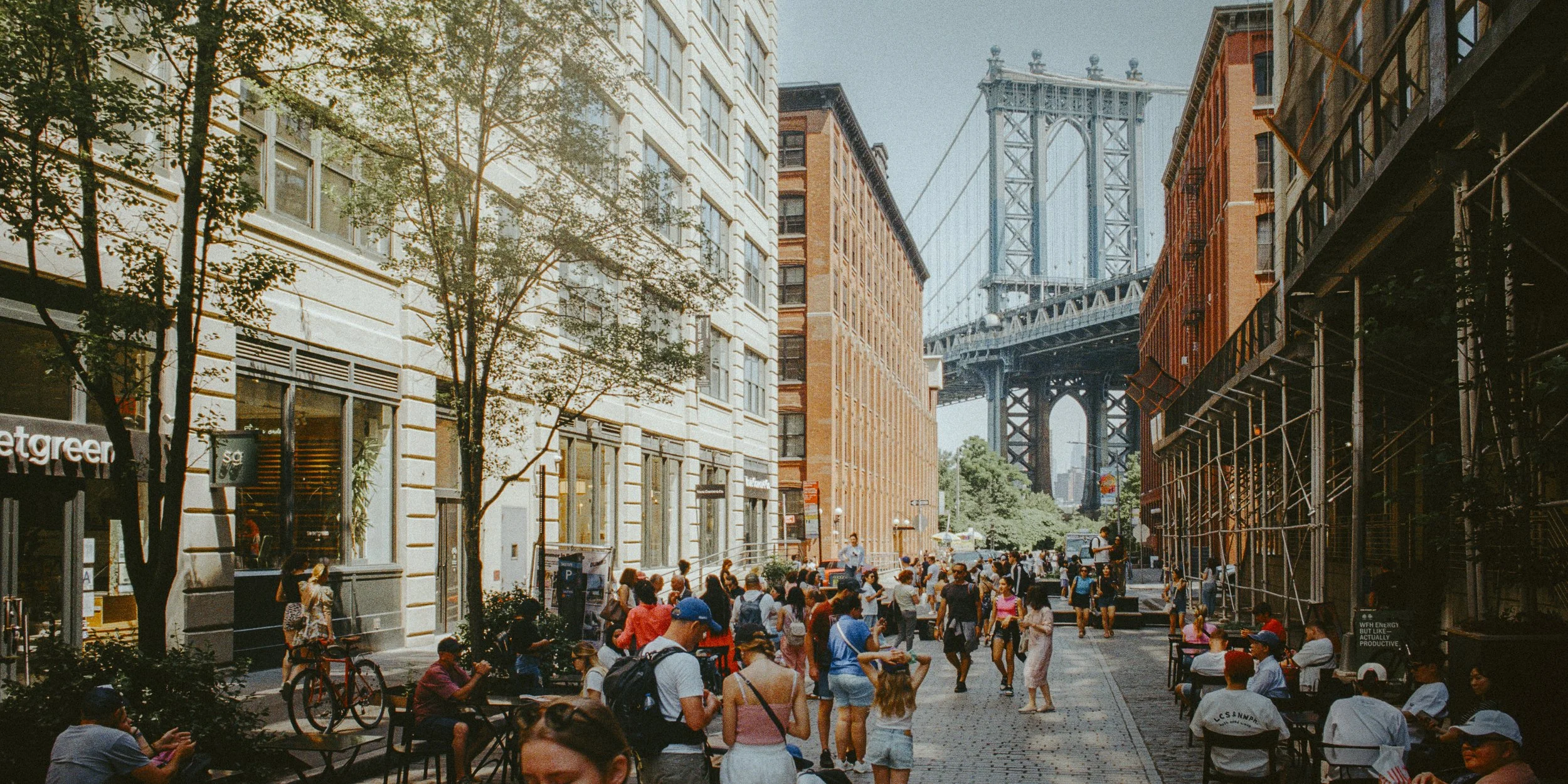 Street scene in New York City with the Brooklyn Bridge in the background, people walking and sitting at outdoor cafes, trees, and urban buildings.