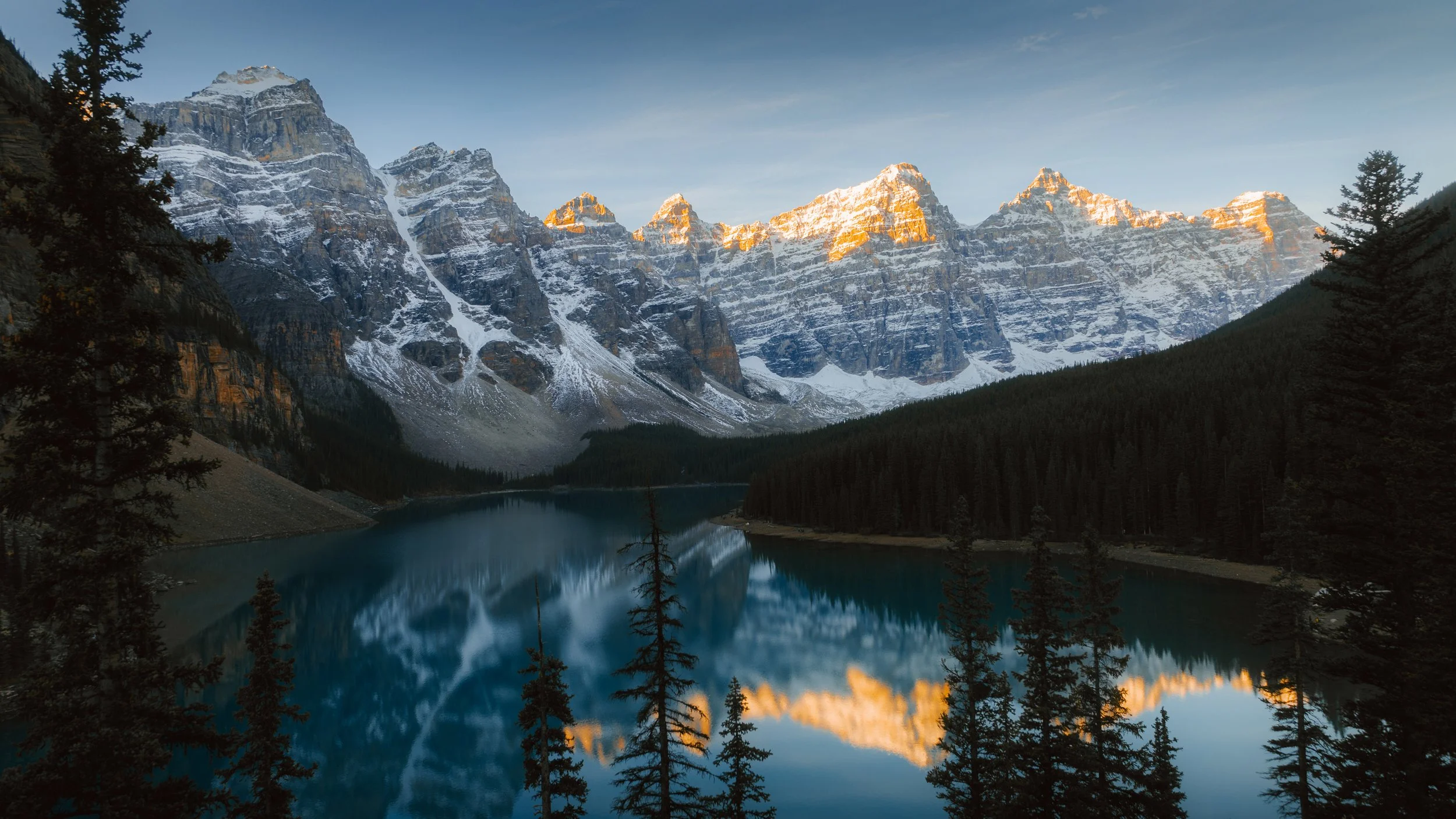 Snow-capped mountains reflecting in a calm lake surrounded by pine trees at sunset.