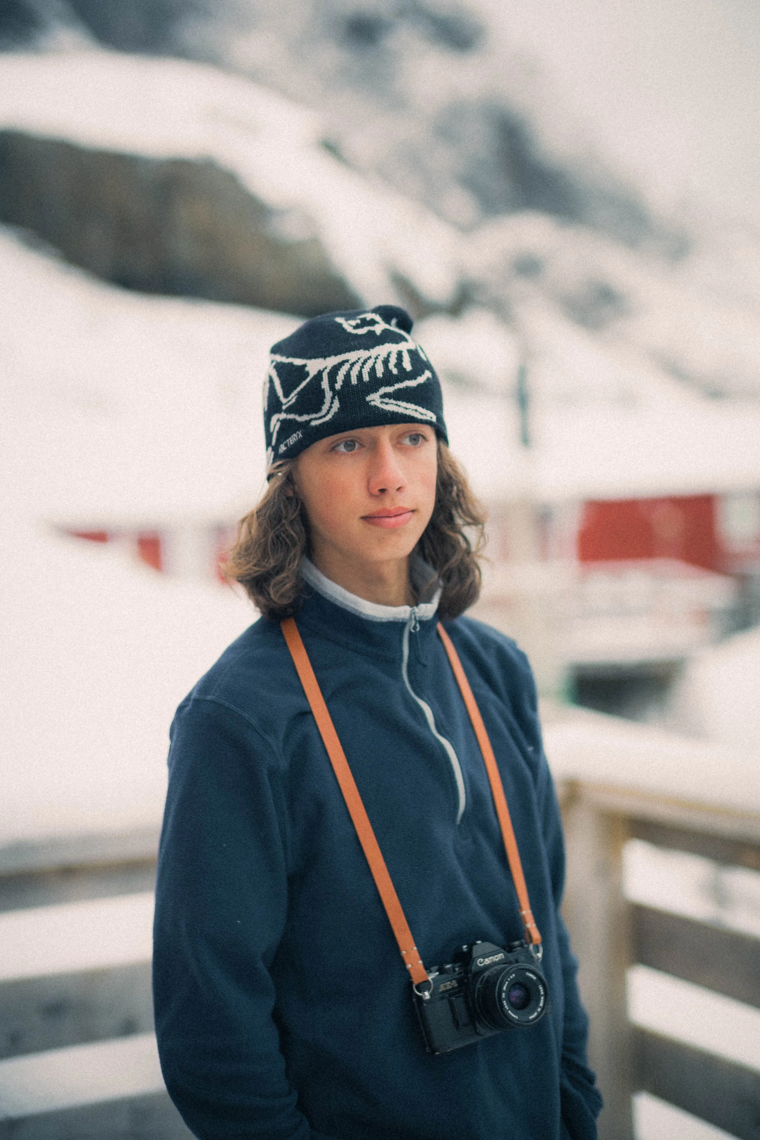 A young person with long hair wearing a black beanie hat with a white skull design, a navy blue zip-up jacket, and a camera hanging around their neck, standing outdoors in a snowy mountain landscape.
