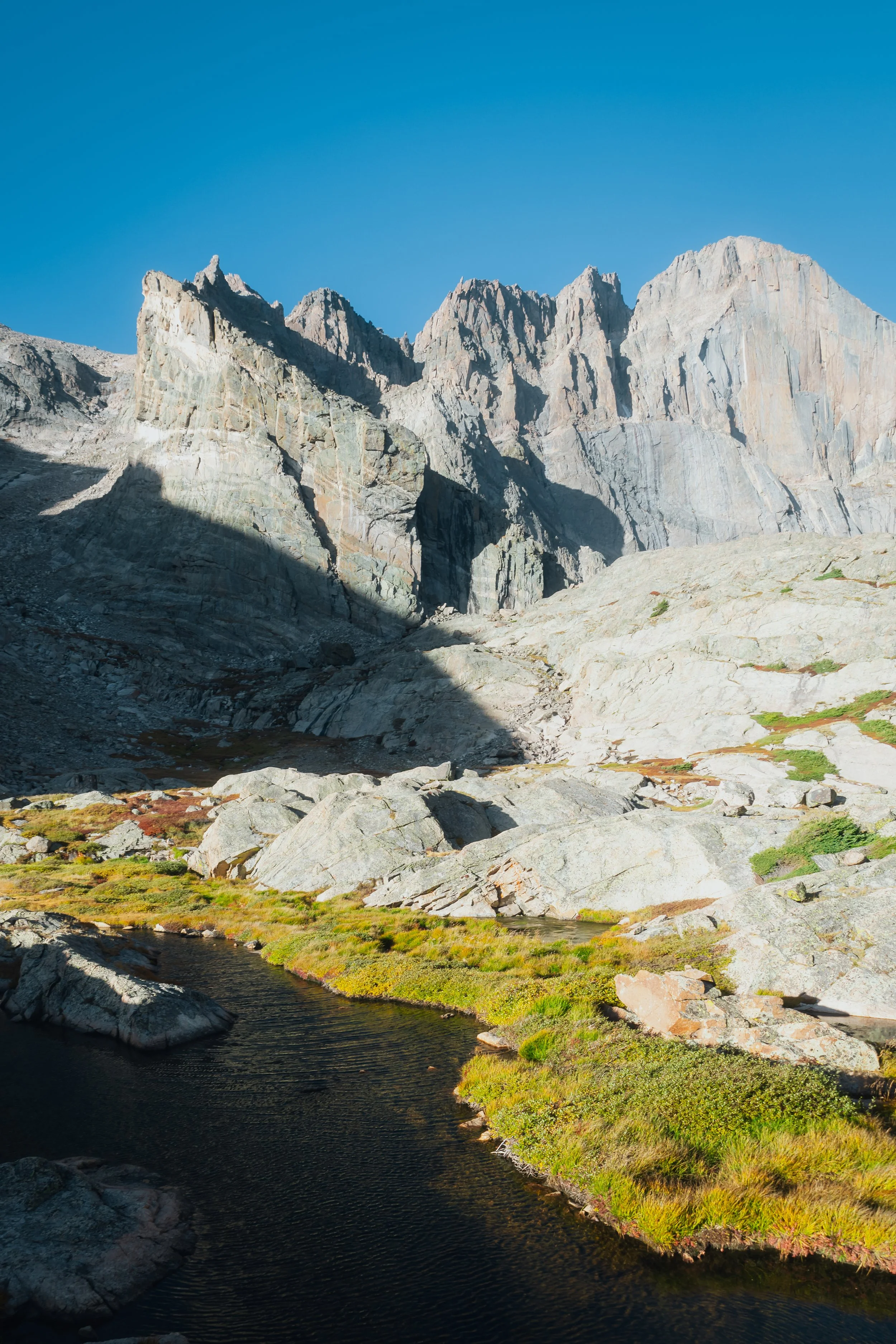 A rugged mountain landscape with tall gray rock formations under a clear blue sky, and a small body of dark water surrounded by green and yellow moss in the foreground.
