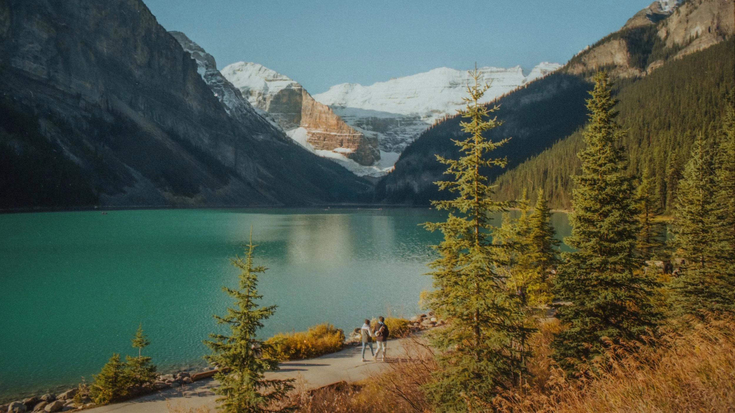Two people walking along a trail beside a turquoise lake surrounded by evergreen trees and snow-capped mountains in the distance.