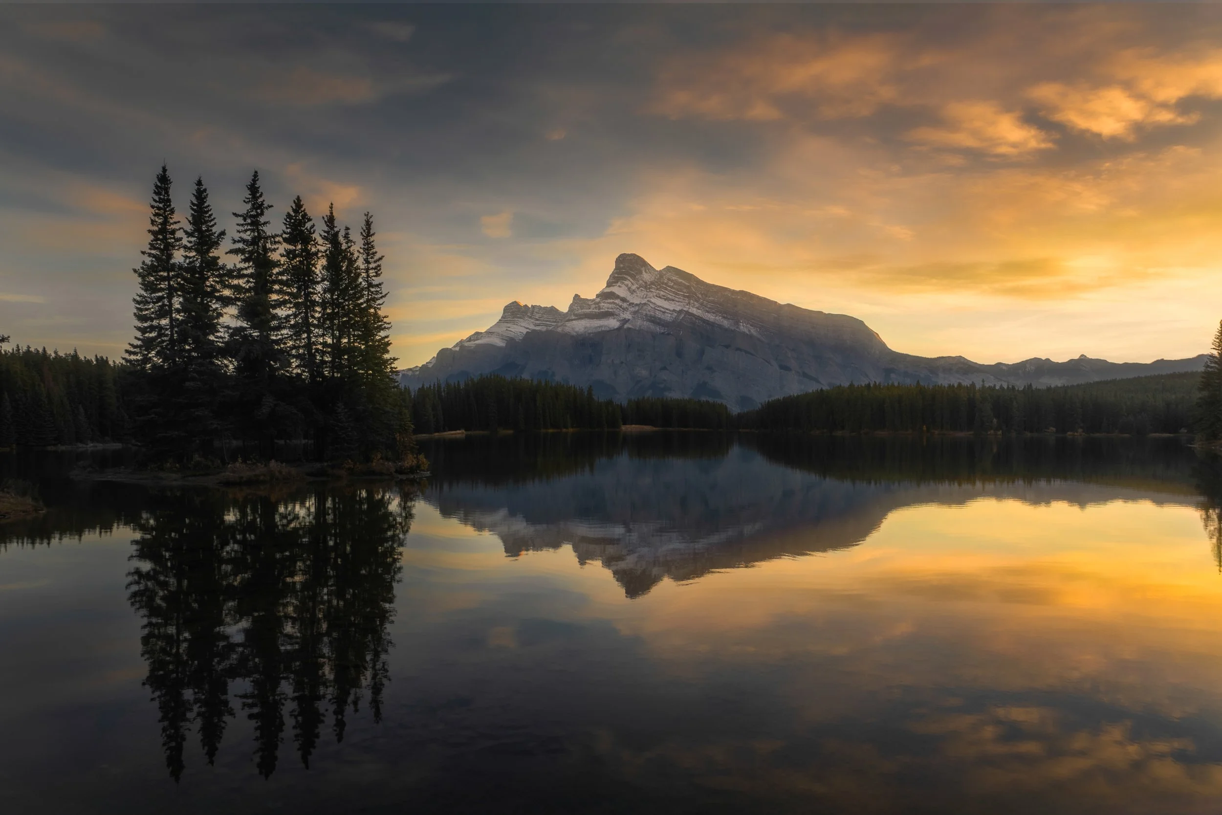 A serene lake reflecting a mountain range and forest, with a sunset sky featuring orange and yellow hues.
