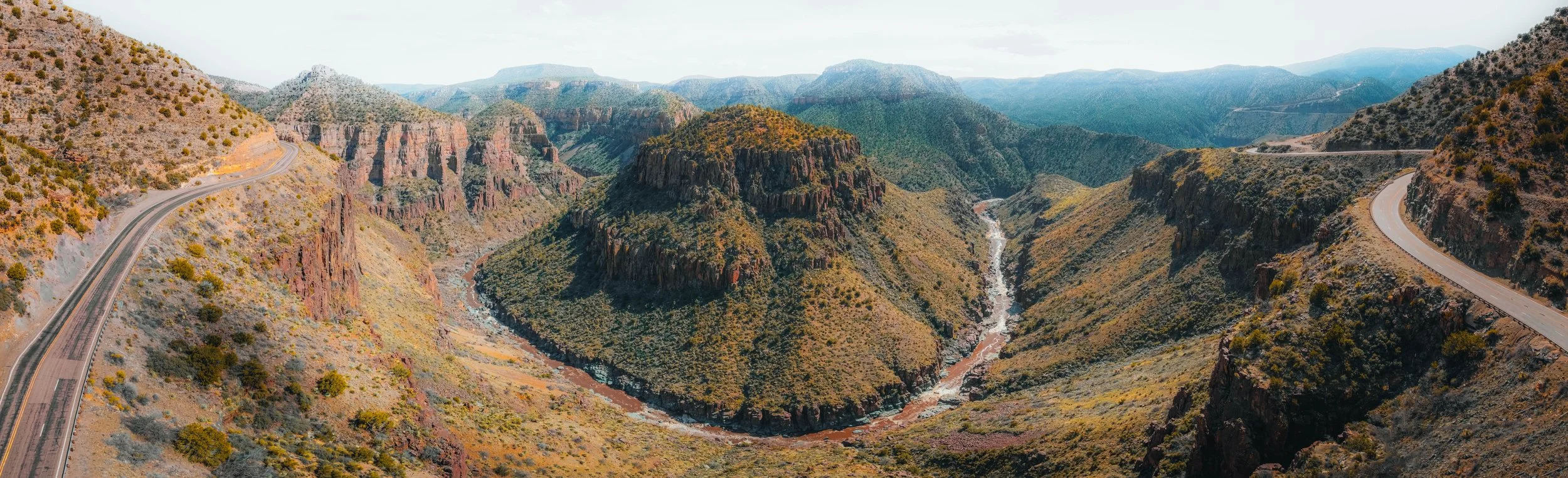 Panoramic view of the Grand Canyon showcasing steep, rugged cliffs, a winding river at the bottom, and a road along the rim on the right side.