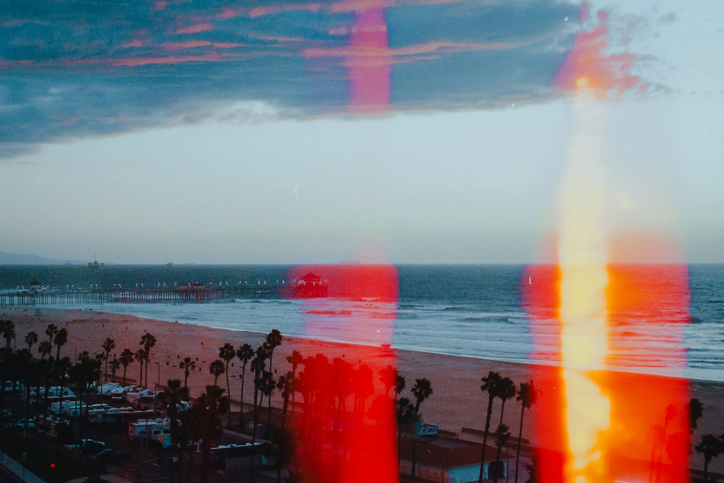 Beach scene with palm trees, a pier extending into the ocean, and a sunset sky with clouds and a rainbow reflection on the water, seen through a window with light leaks.