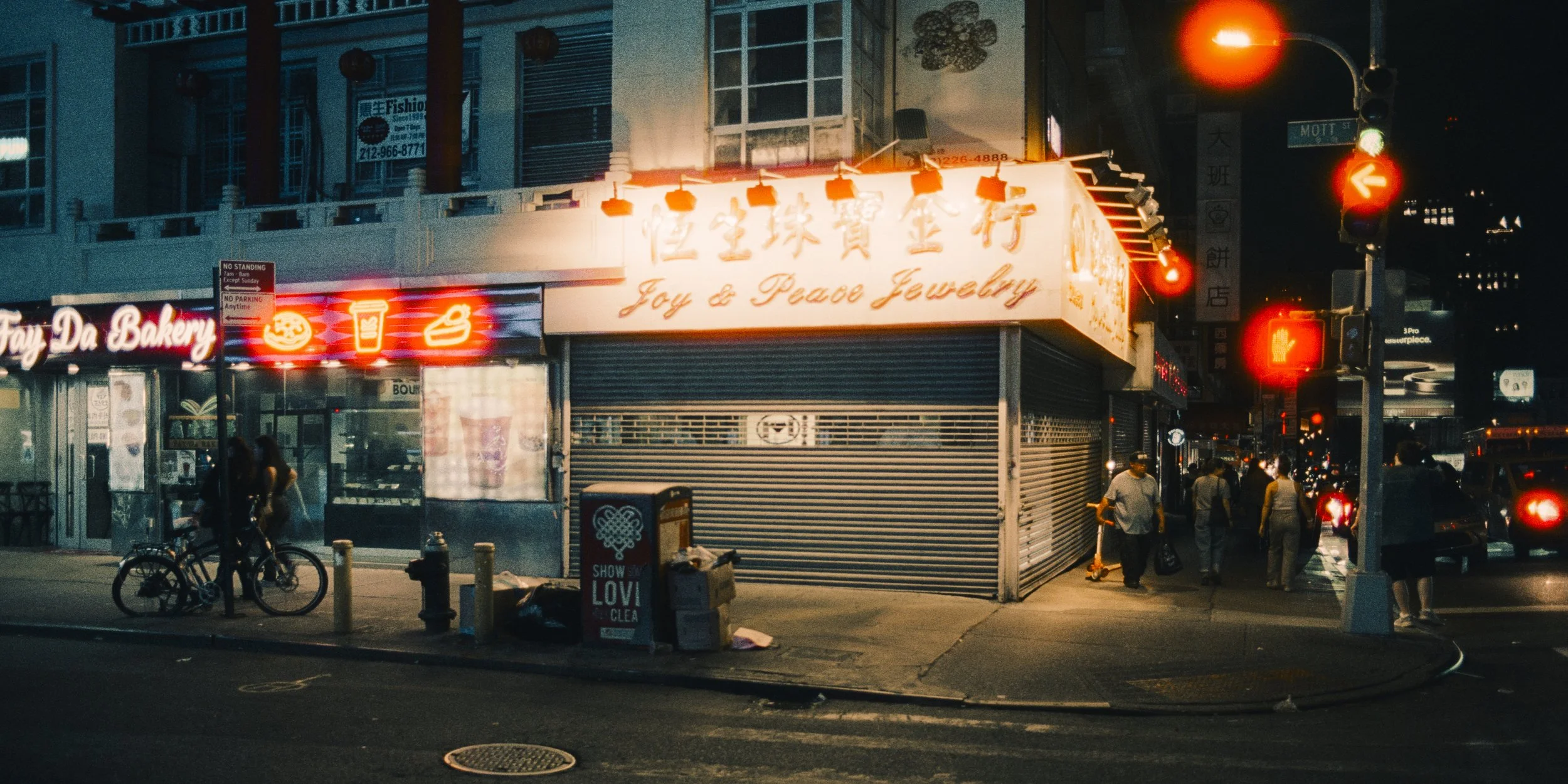 Nighttime street scene with a closed jewelry store sign reading 'Joy & Peace Jewelry' and a bakery with neon signs, including a pumpkin, a drink, and a cone. People walk on the sidewalk, some on bicycles, with city traffic and illuminated traffic lig