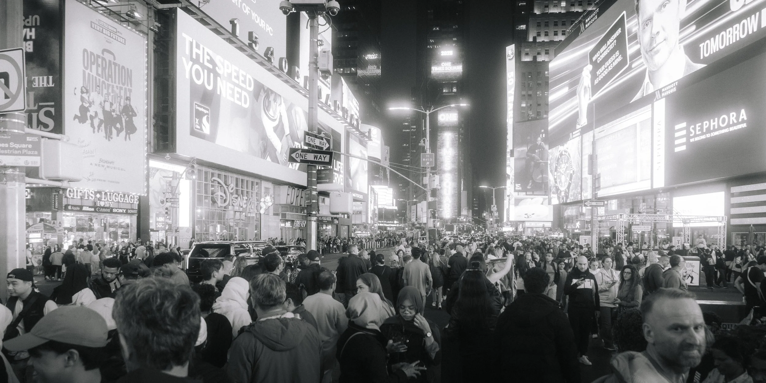 Crowded city street at night with bright neon billboards and digital screens, showing advertisements and messages, amidst a large group of pedestrians.