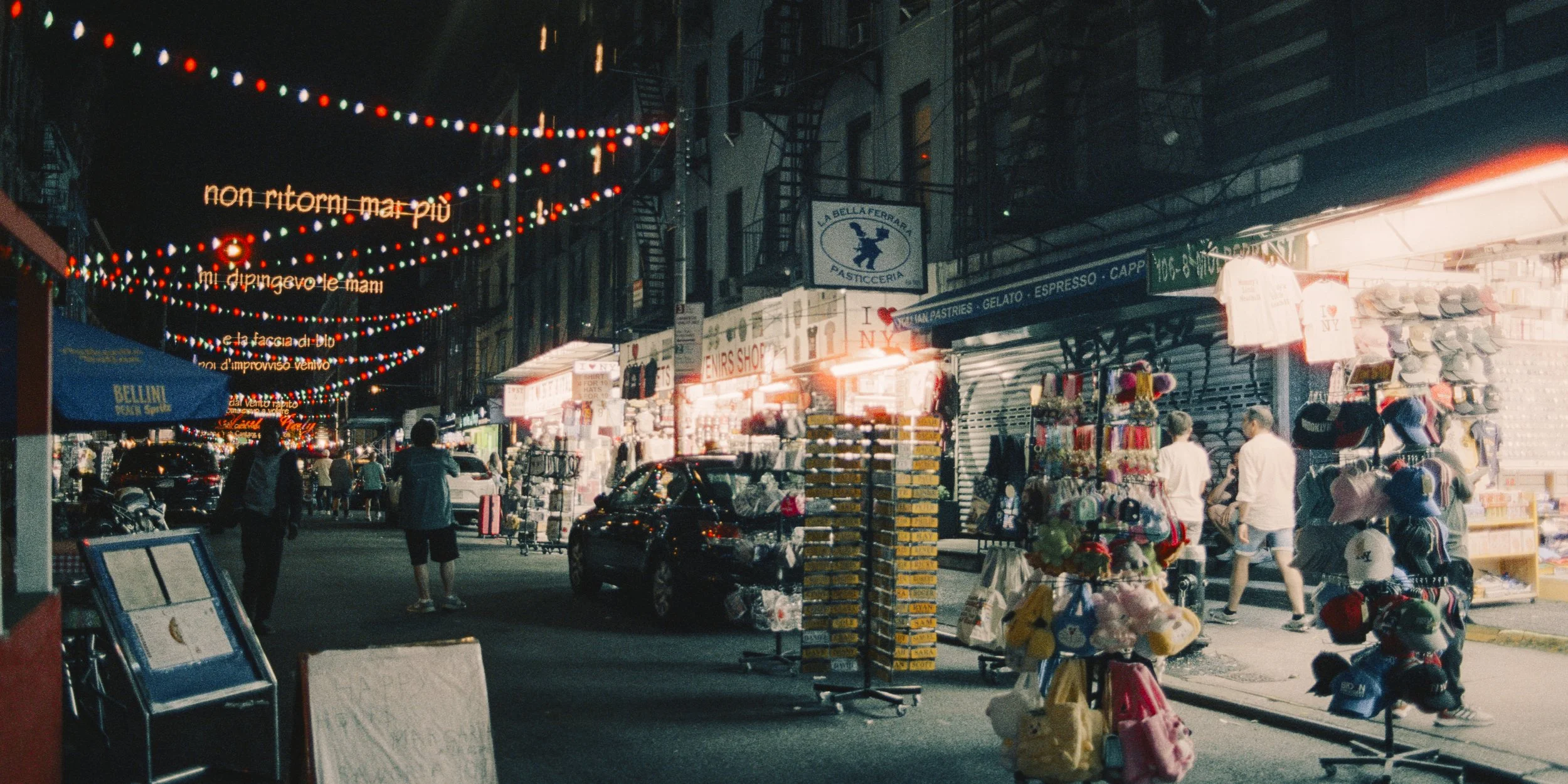 Street scene at night with shops selling hats, bags, and souvenirs, illuminated by streetlights and hanging string lights. People are walking along the sidewalk.