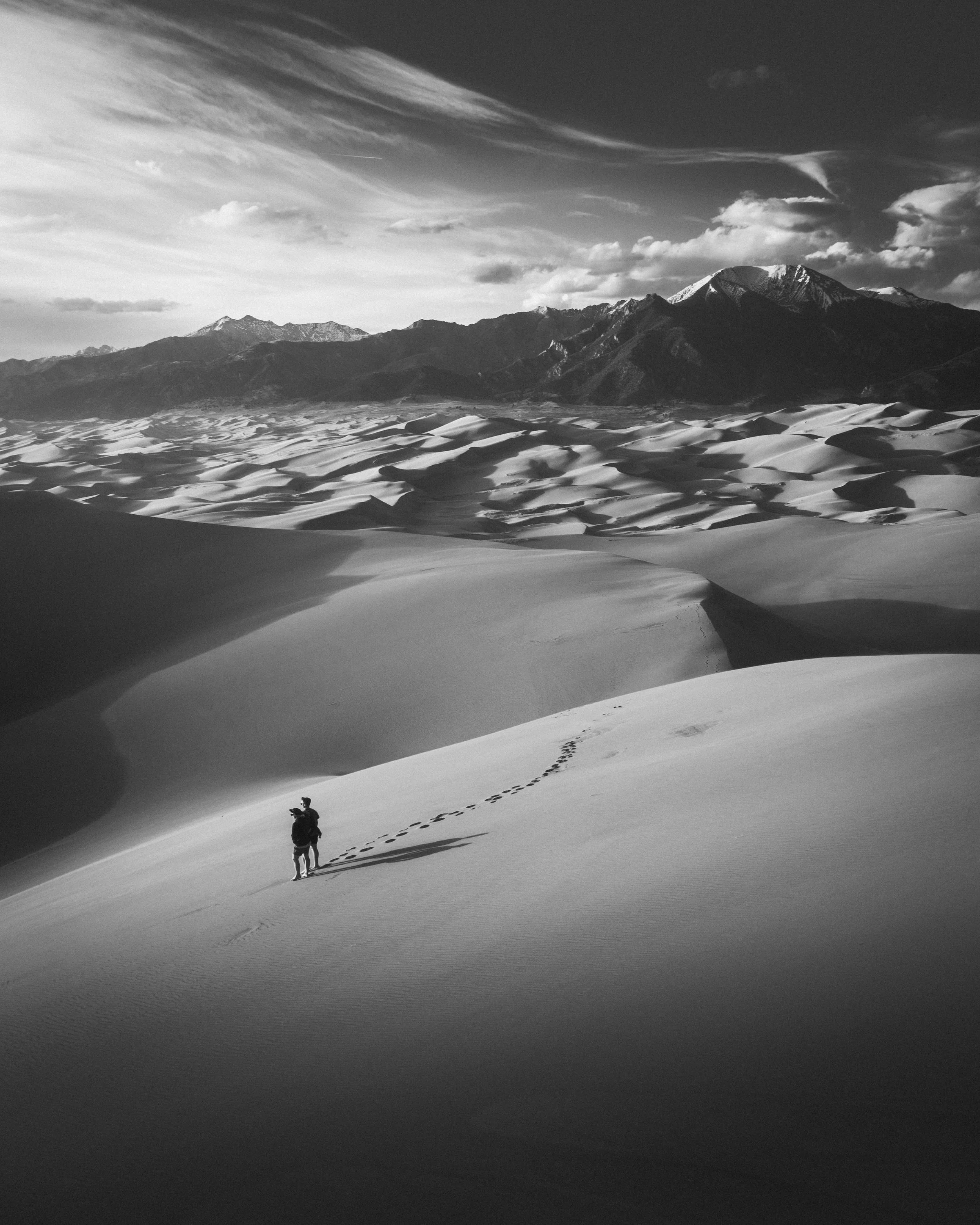 A person standing in a vast desert with sand dunes, mountains in the background, and a cloudy sky overhead.