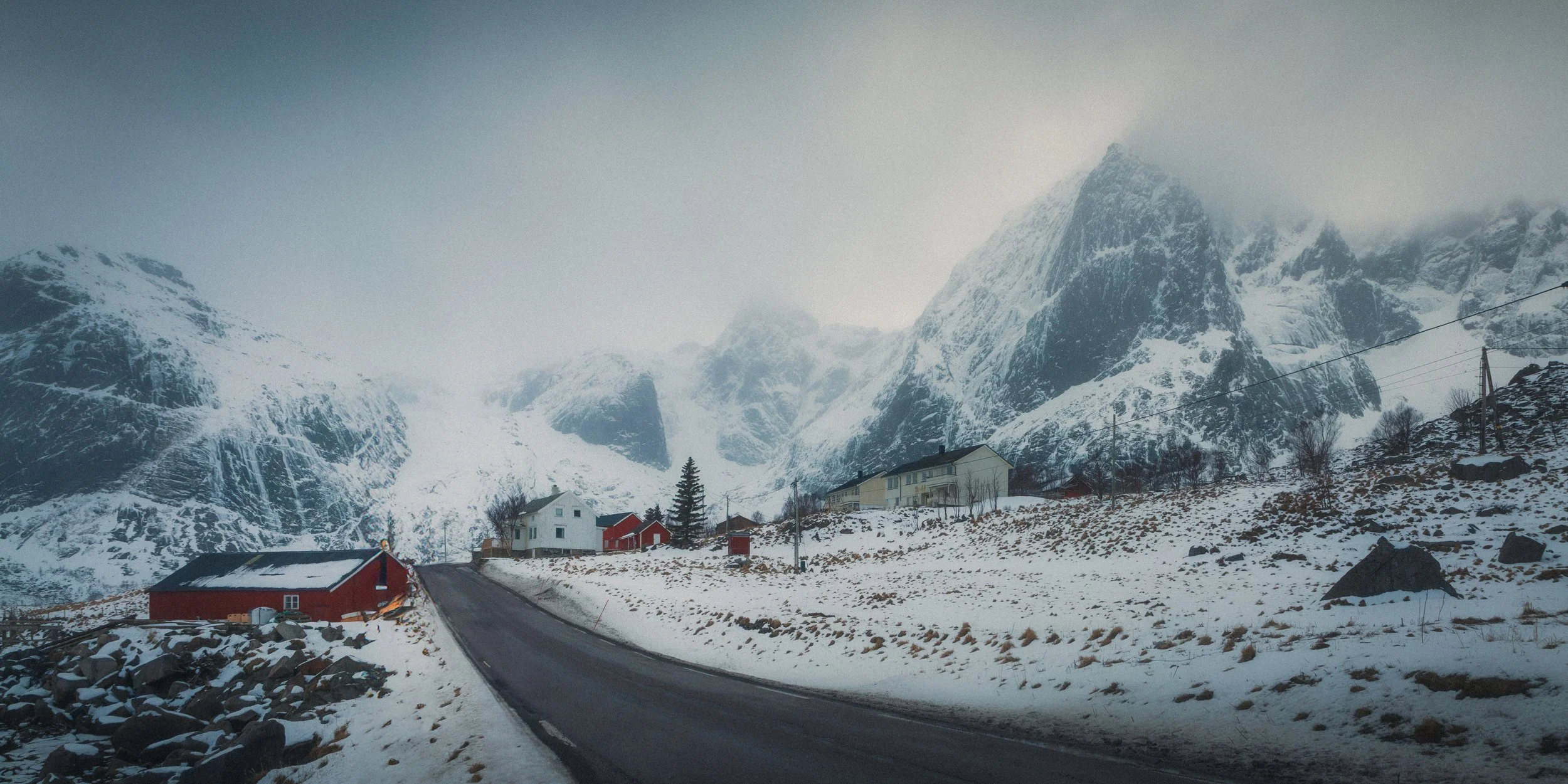 Snow-covered mountain landscape with a winding road, scattered houses, and trees in a cold, foggy environment.