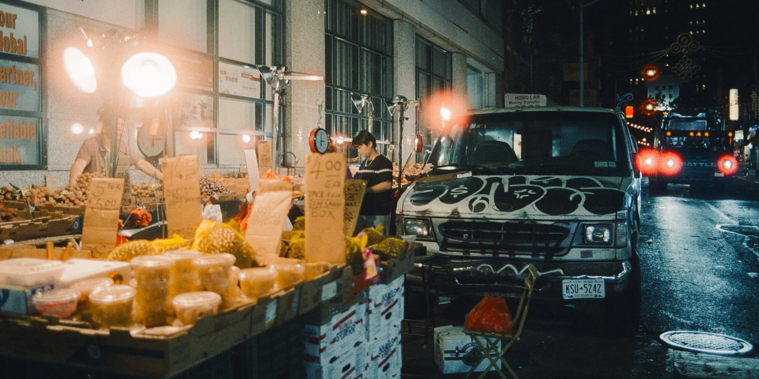 Night scene of a street food stall with a variety of fruits and packaged goods on display, illuminated by bright lights. A person is browsing the items, with cars parked nearby and city lights in the background.