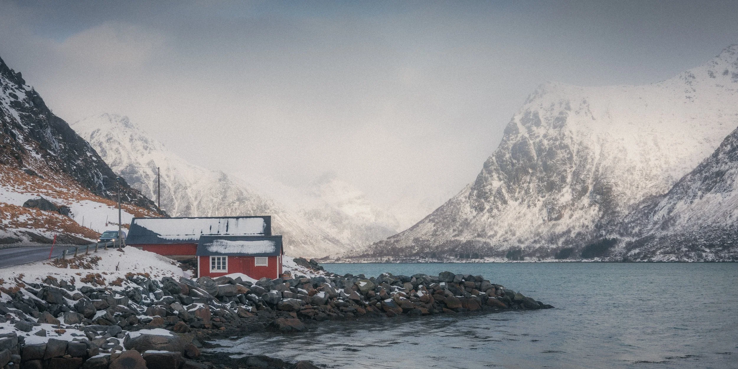 Snow-covered mountains surrounding a fjord with a small red house and a car on the road nearby.