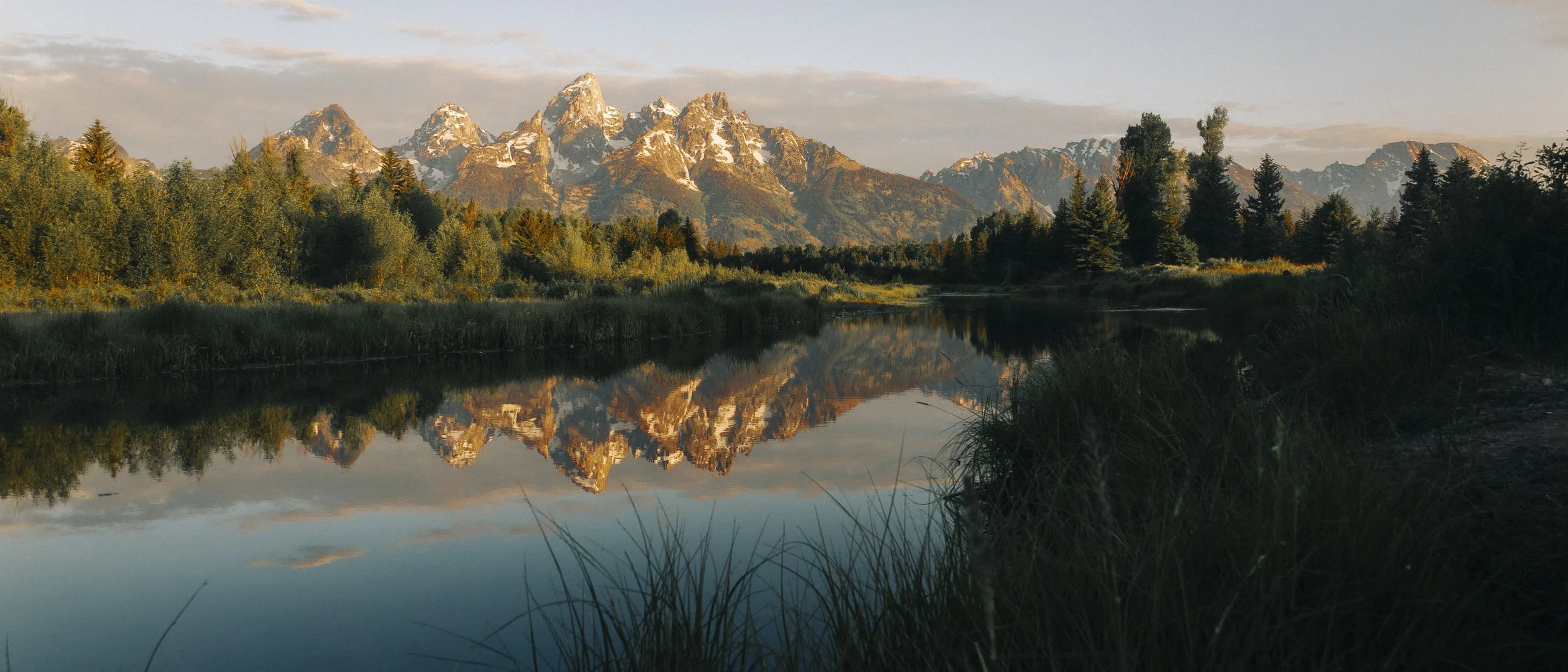 Mountains with snow caps reflected in a calm river, surrounded by lush green trees and grass during sunset or sunrise.