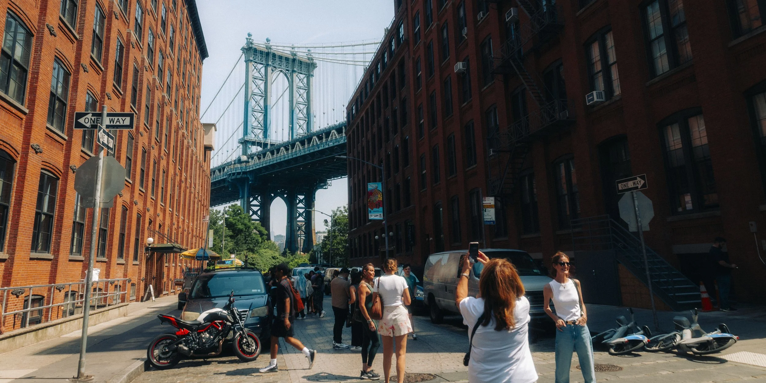 People walking and taking photos on a city street near the Manhattan Bridge in New York City, with brick buildings on each side and parked motorcycles and scooters.