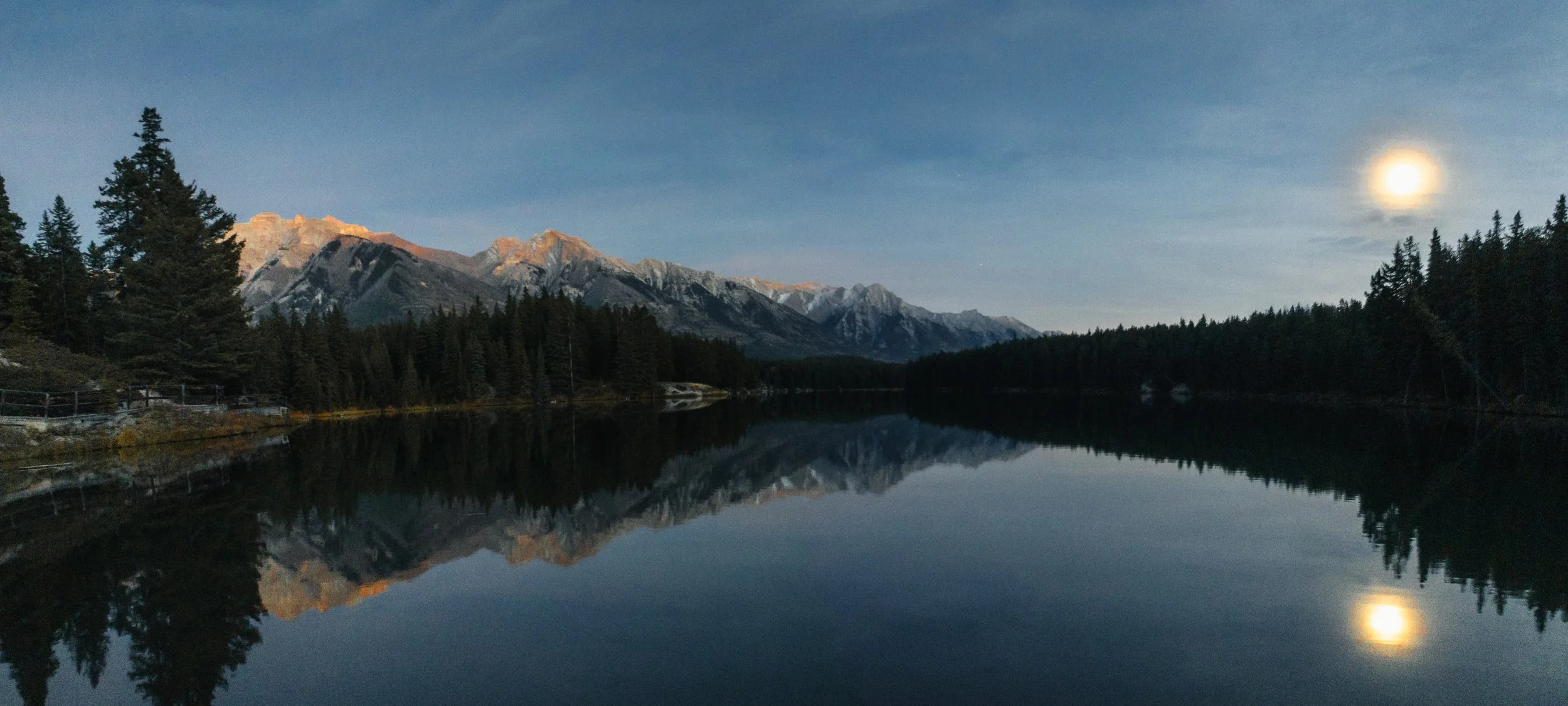 A tranquil lake reflecting a forested shoreline, snow-capped mountains in the background, and a bright moon in the night sky.