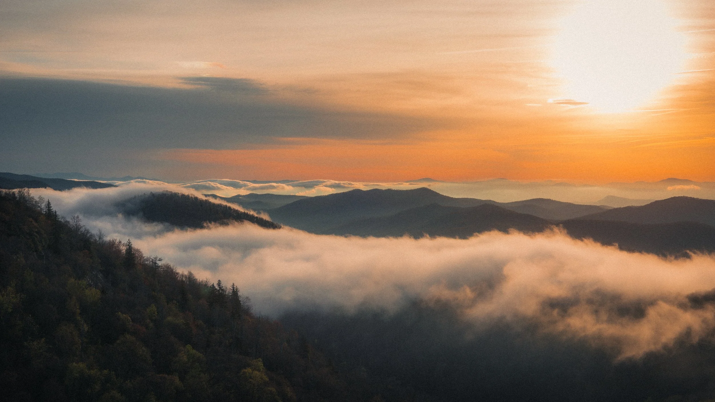 Sunset over misty mountain ranges with clouds and fog in a valley, hues of orange and pink in sky.