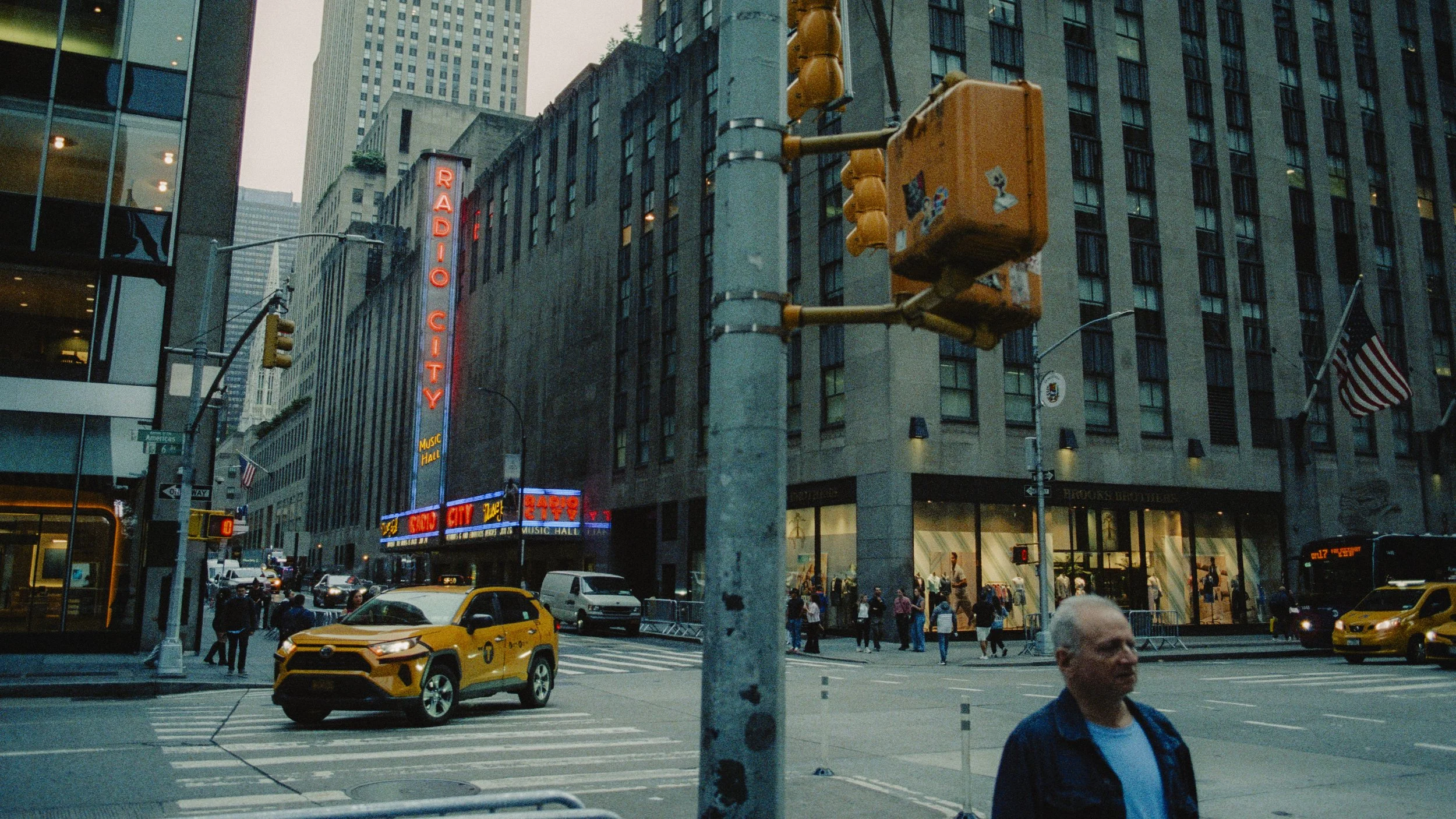 A city street intersection with a yellow taxi, pedestrians, and tall buildings. A neon sign reads 'Radio City Music Hall' on one building, and an American flag is displayed. Traffic lights and street signs are visible.