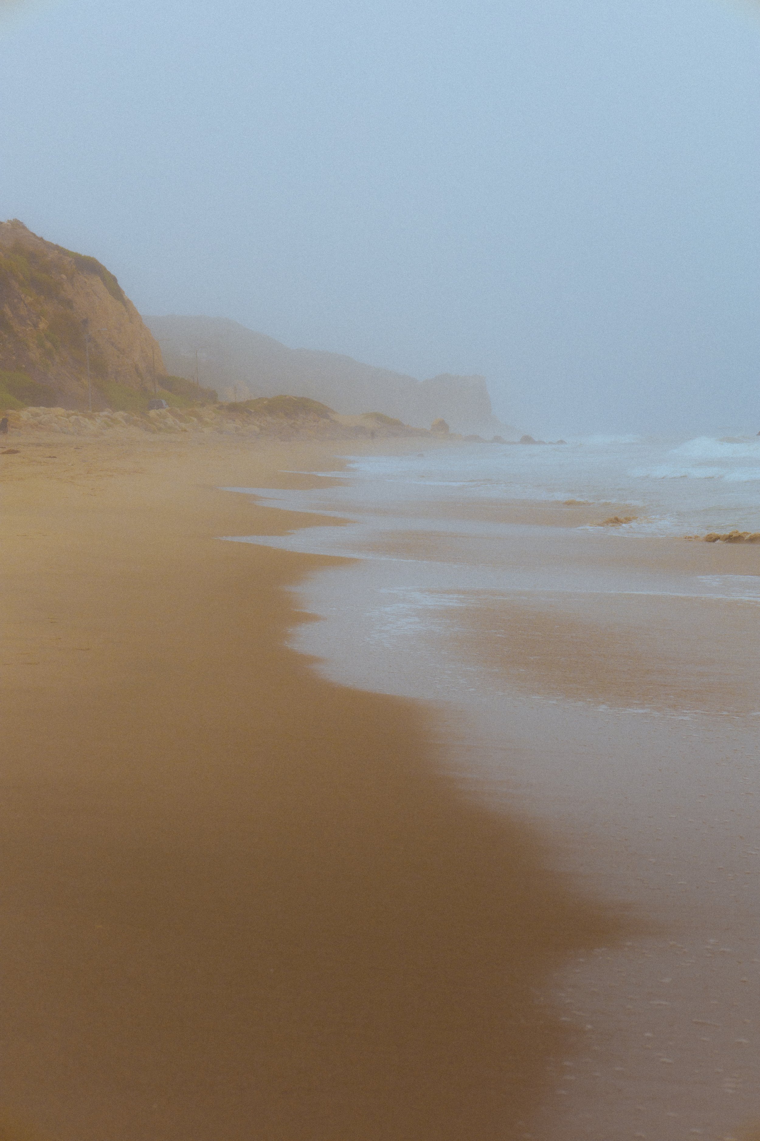 A foggy beach scene with sandy shore, ocean waves, and cliffs in the background.