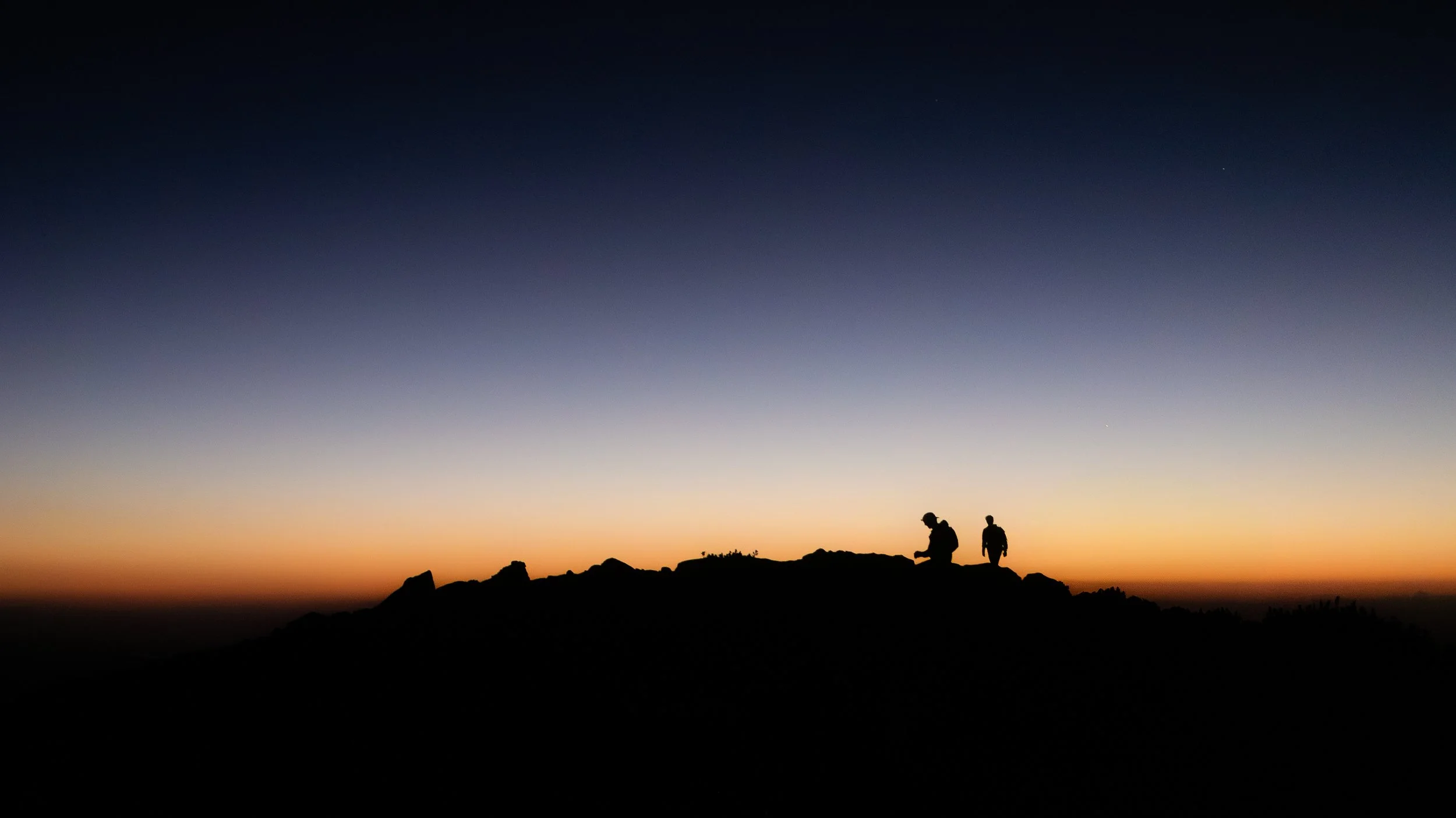 Silhouettes of two hikers on a mountain ridge during sunset with a colorful sky in background.