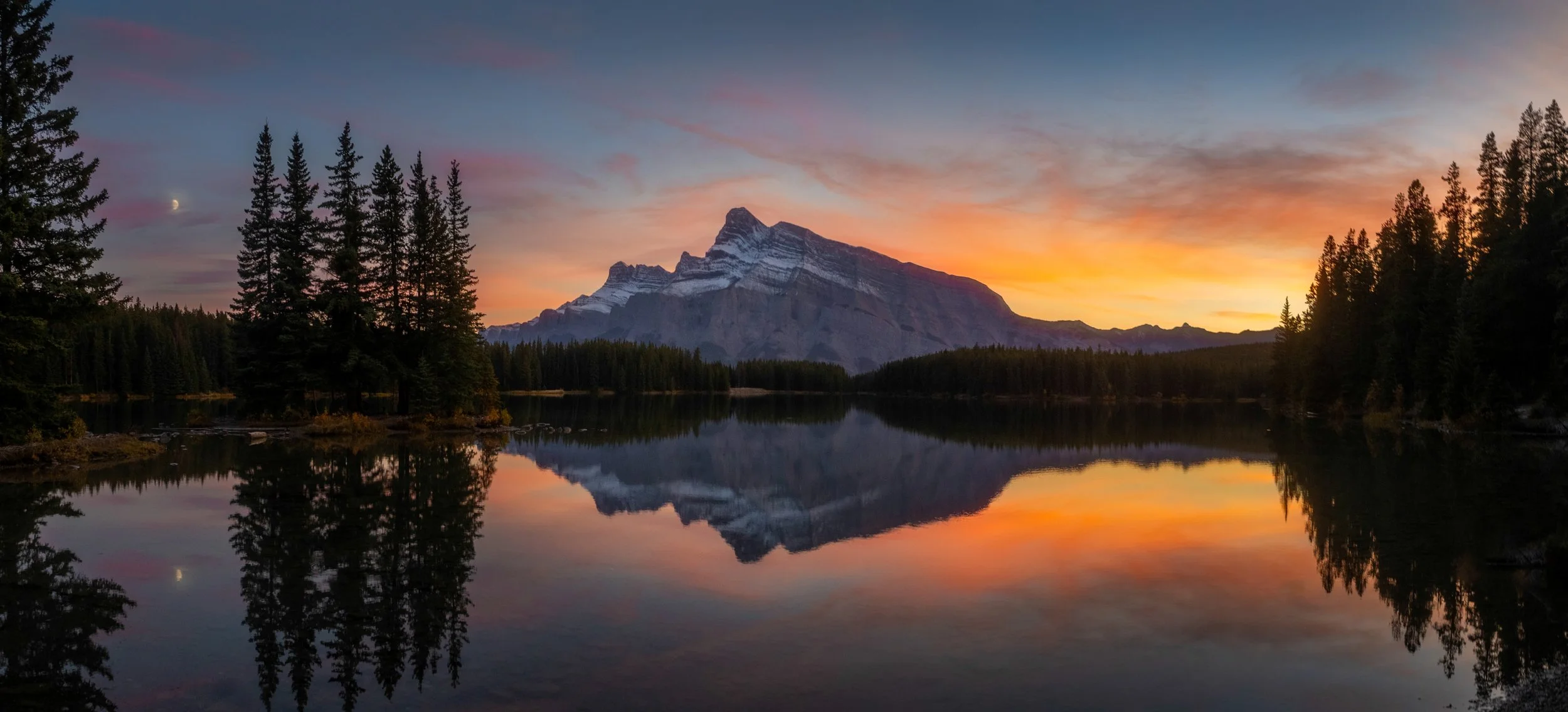 A serene landscape of a mountain with snow patches, surrounded by tall pine trees, reflected in a calm lake during sunset with a colorful sky.