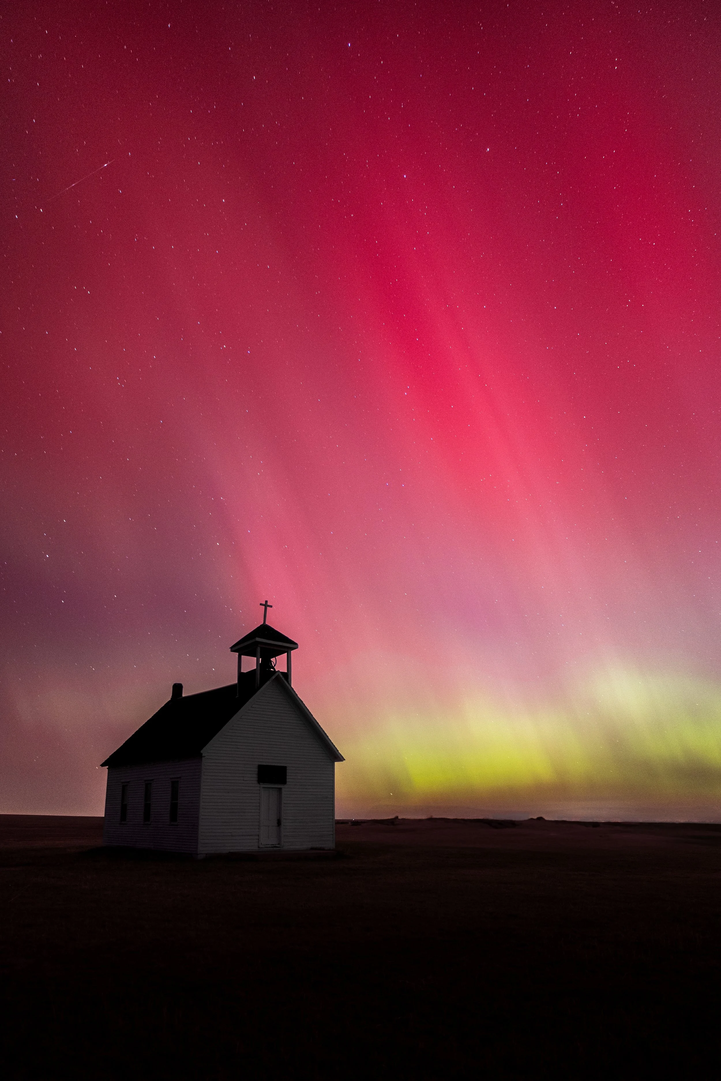 A small white church building with a steeple and cross is silhouetted against a colorful night sky with the aurora borealis and stars.