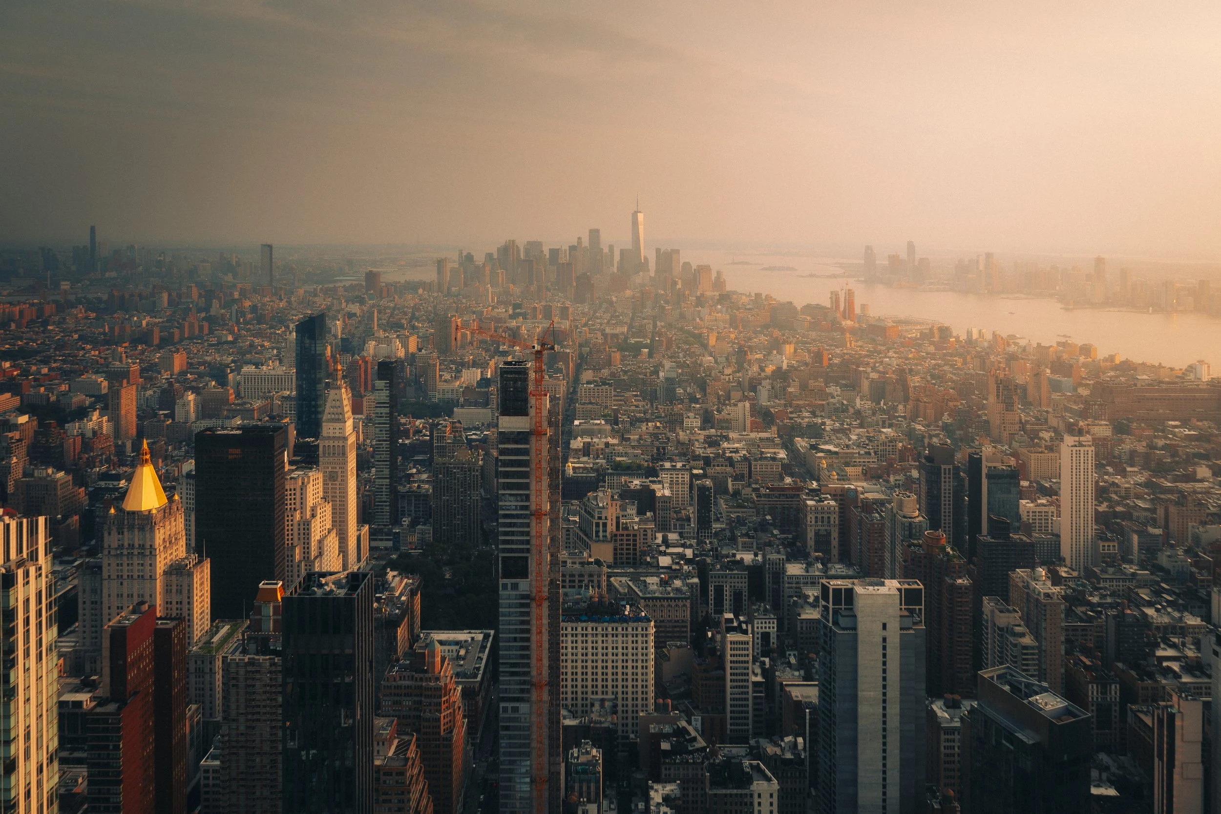 Aerial view of New York City skyline at sunset, with numerous skyscrapers, including One World Trade Center, and the Hudson River in the background.
