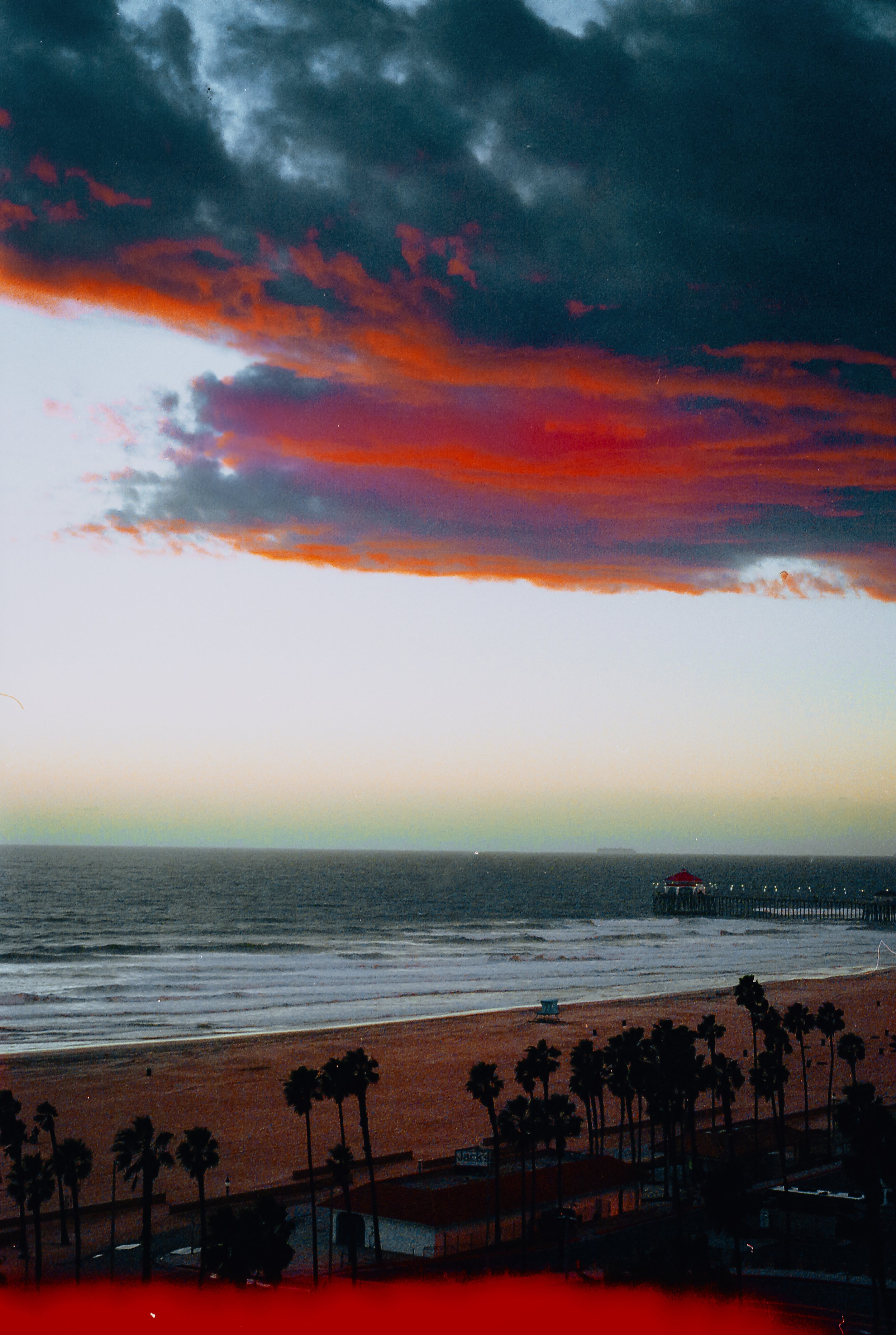 Sunset over the ocean with colorful clouds, beach with palm trees, and a pier extending into the water.