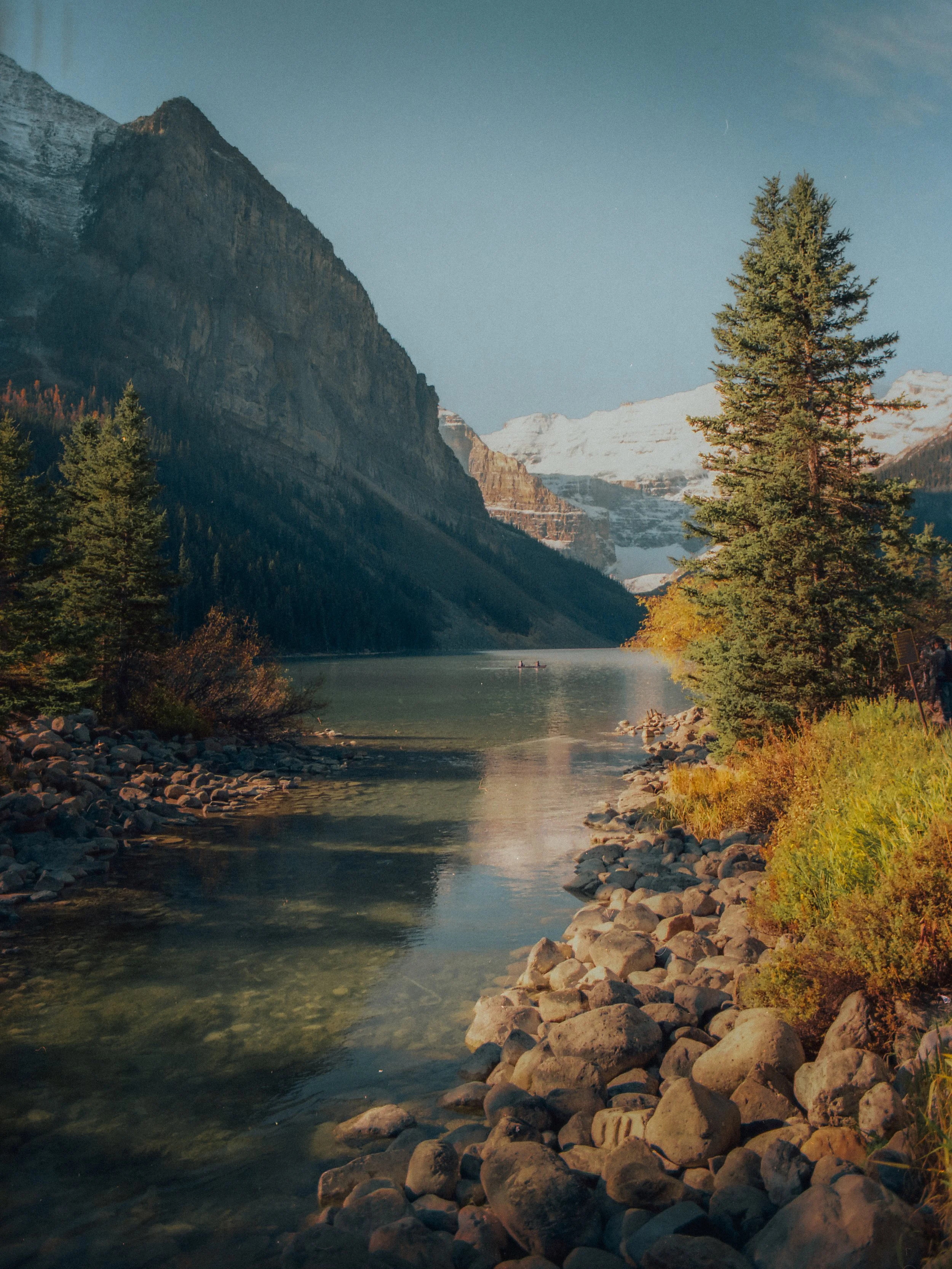 Scenic view of a river with rocks along the banks, surrounded by pine trees, mountains, and snow-capped peaks in the distance under a clear blue sky.