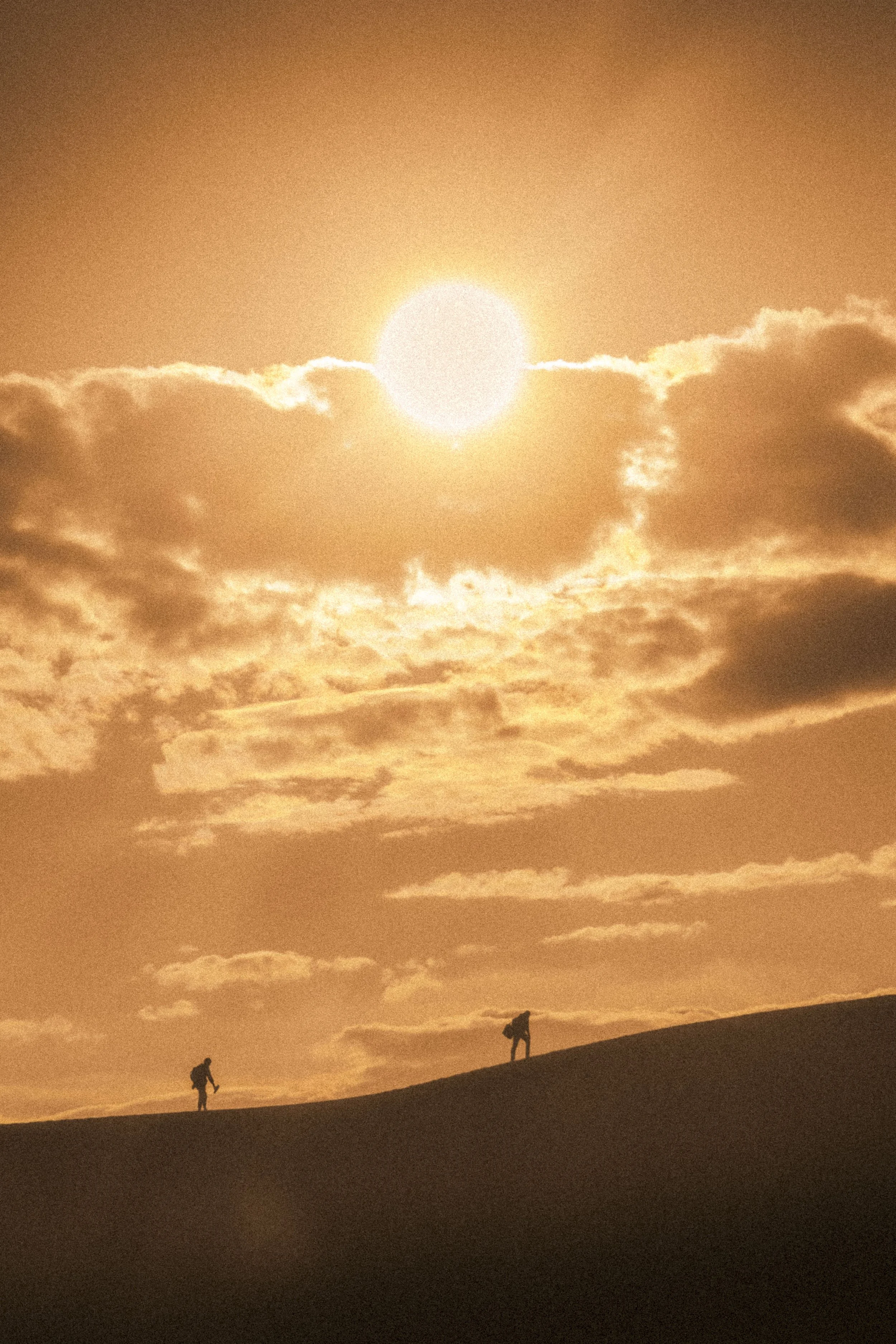 Two hikers with backpacks walking on a sand dune during sunset or sunrise, with a partially cloudy sky and the sun visible near the horizon.