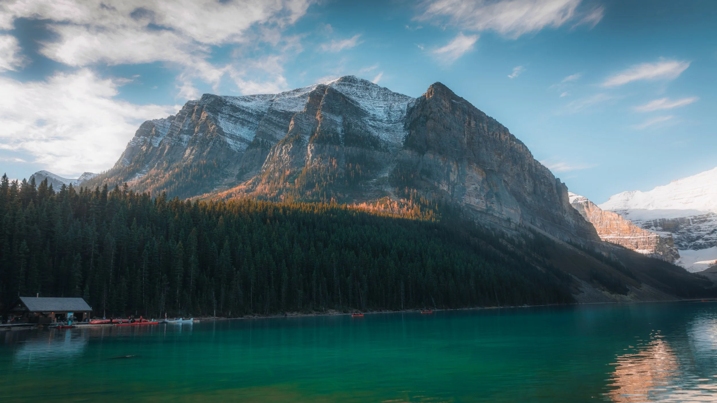 Scenic view of a mountain with snow on top, dense green forest, and a calm teal lake in the foreground, with a wooden cabin on the lake's edge.